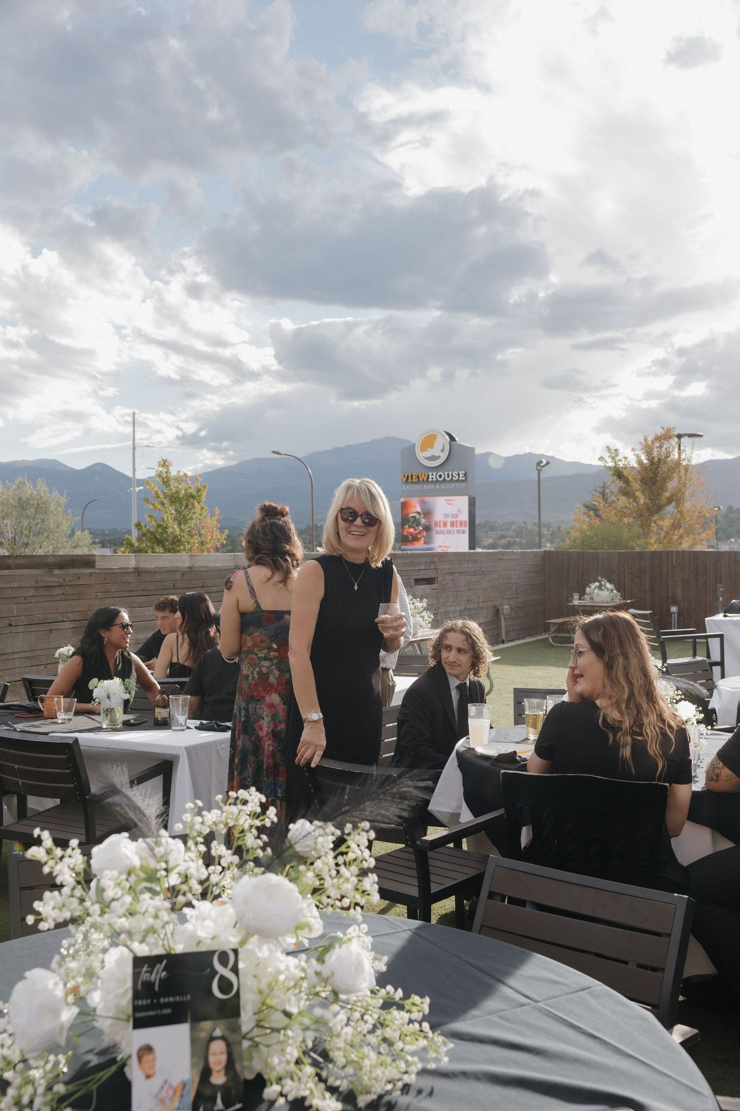 Wedding reception guests mingle on outdoor patio with mountain views and white floral centerpieces.