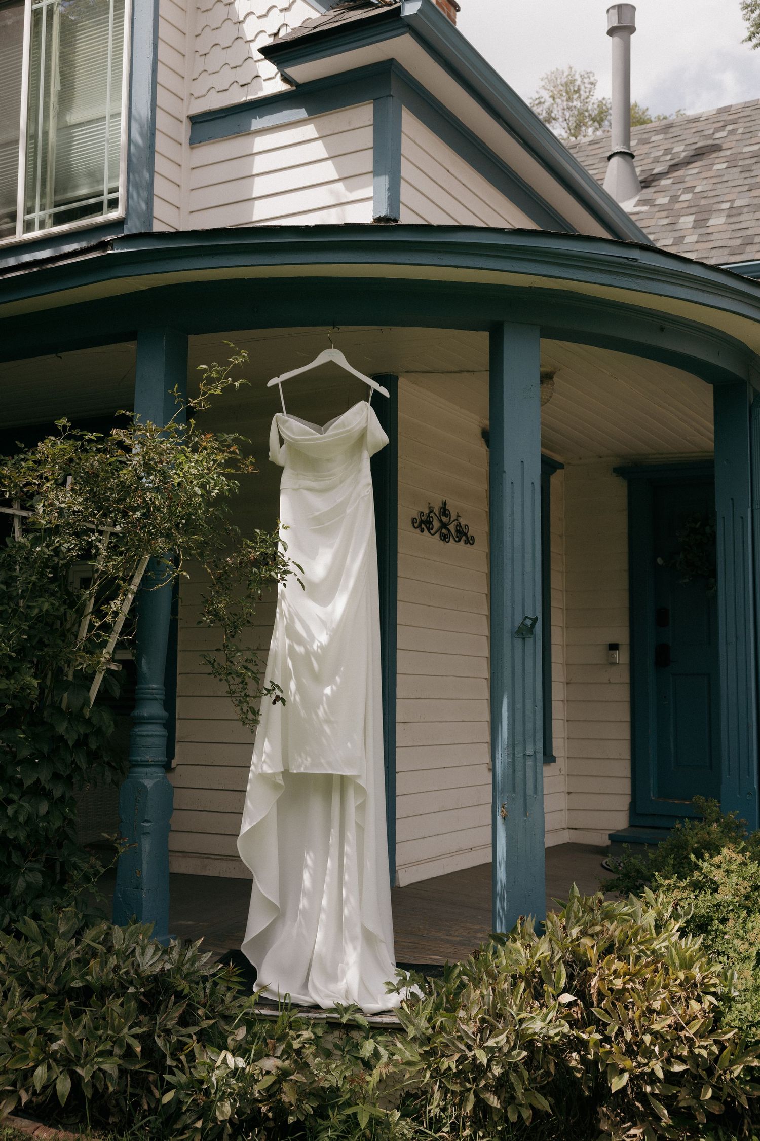 White wedding dress hanging on a hanger on a Victorian porch surrounded by lush greenery.