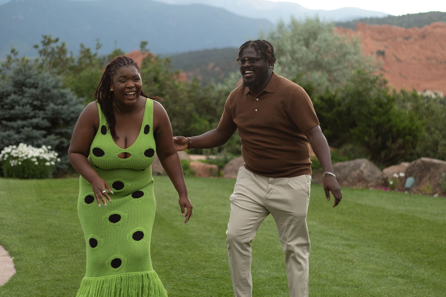A couple laughs and dances together on a green lawn with mountainous scenery in the background.
