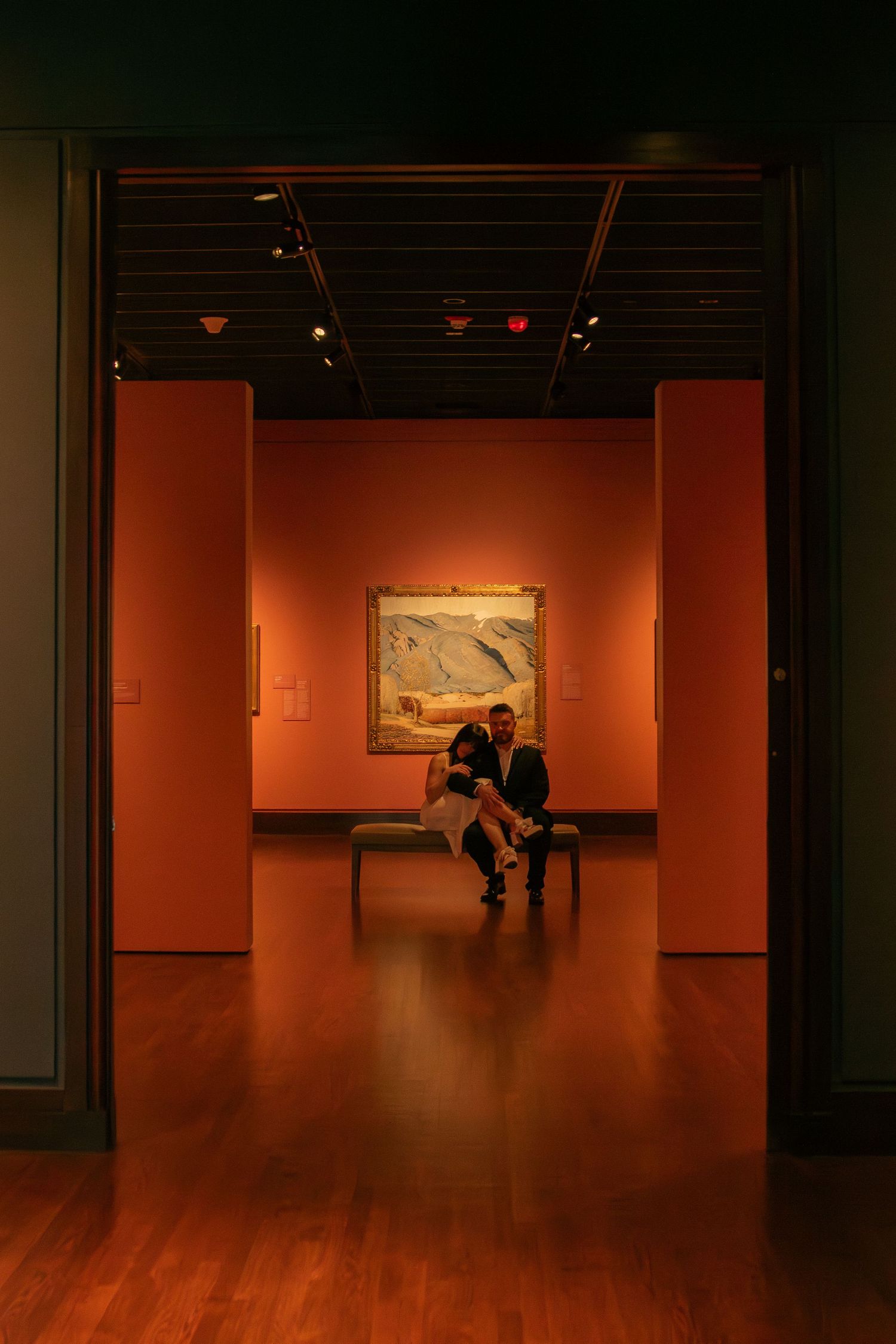 Silhouetted figures in an art gallery with orange walls and wooden floors.