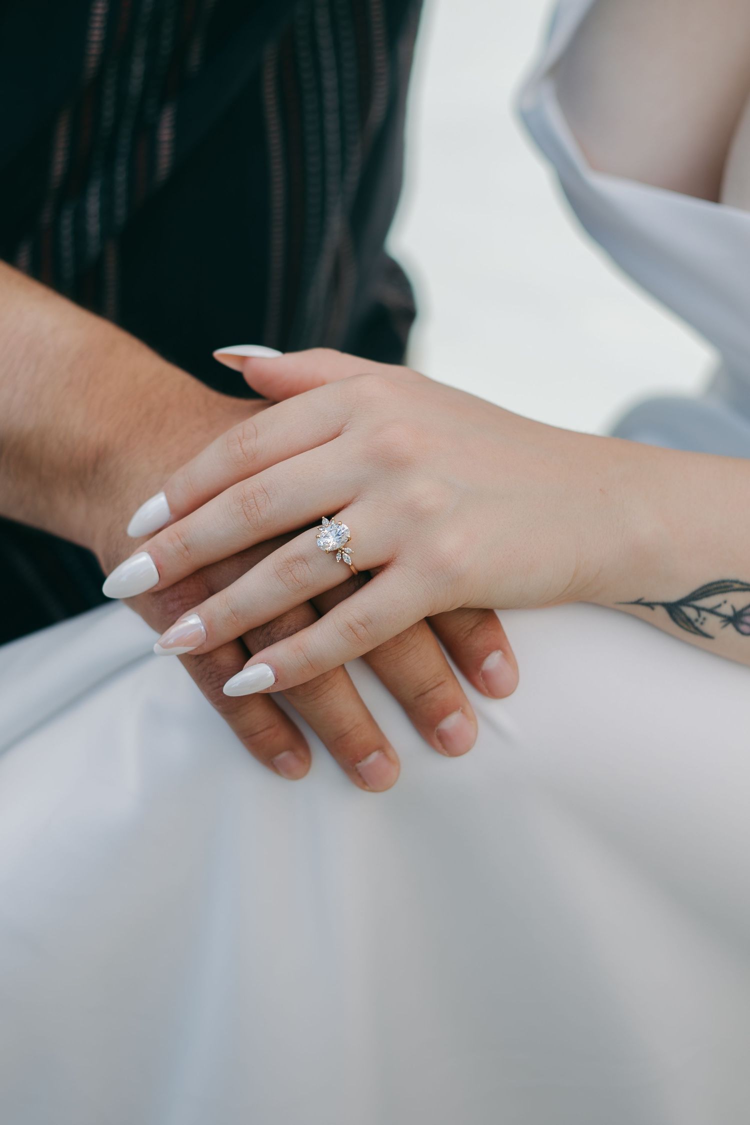 Close-up of intertwined hands with white nail polish and visible tattoo details against light background.