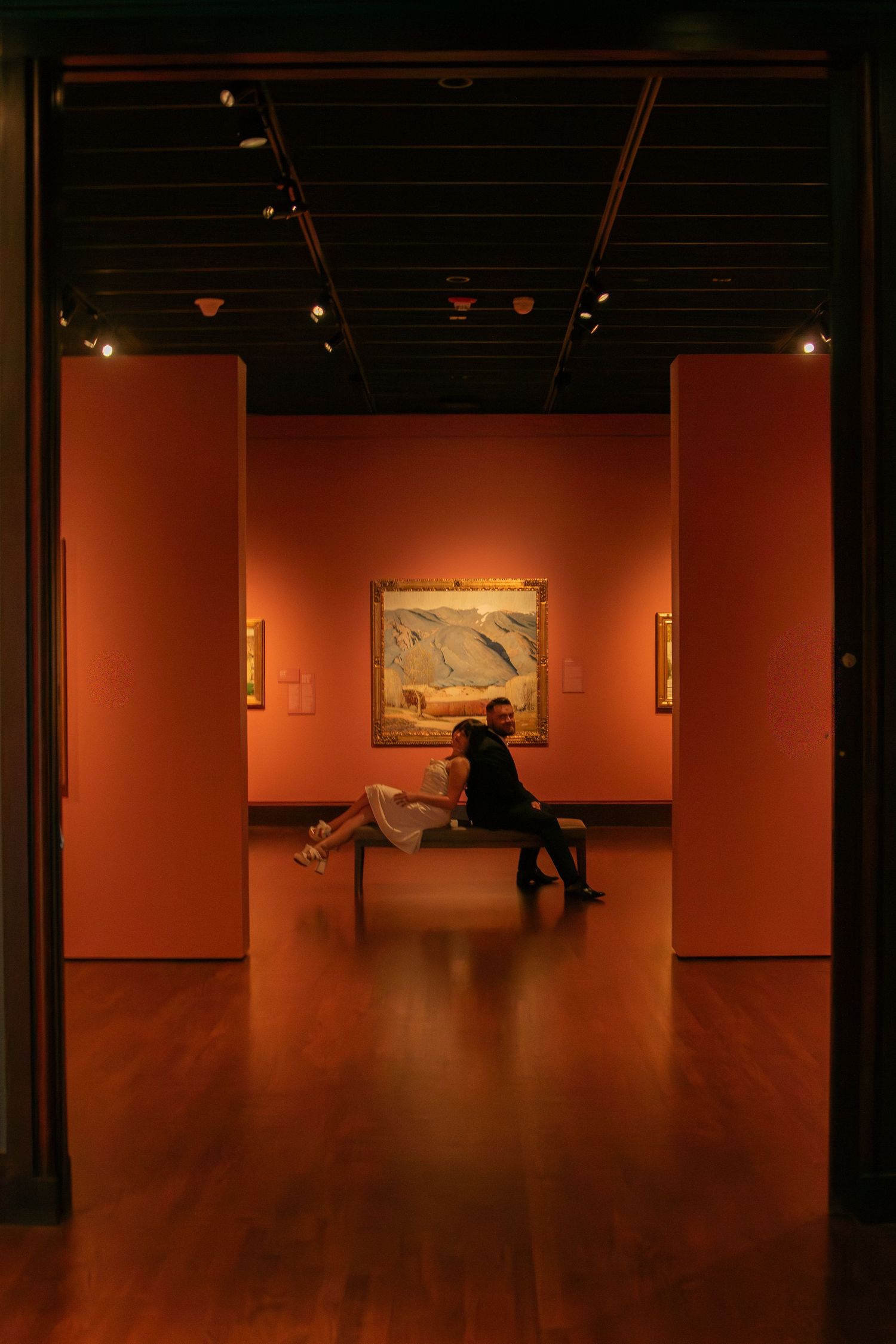 Red-walled art gallery interior with spotlit paintings and distant visitors viewing the exhibits.