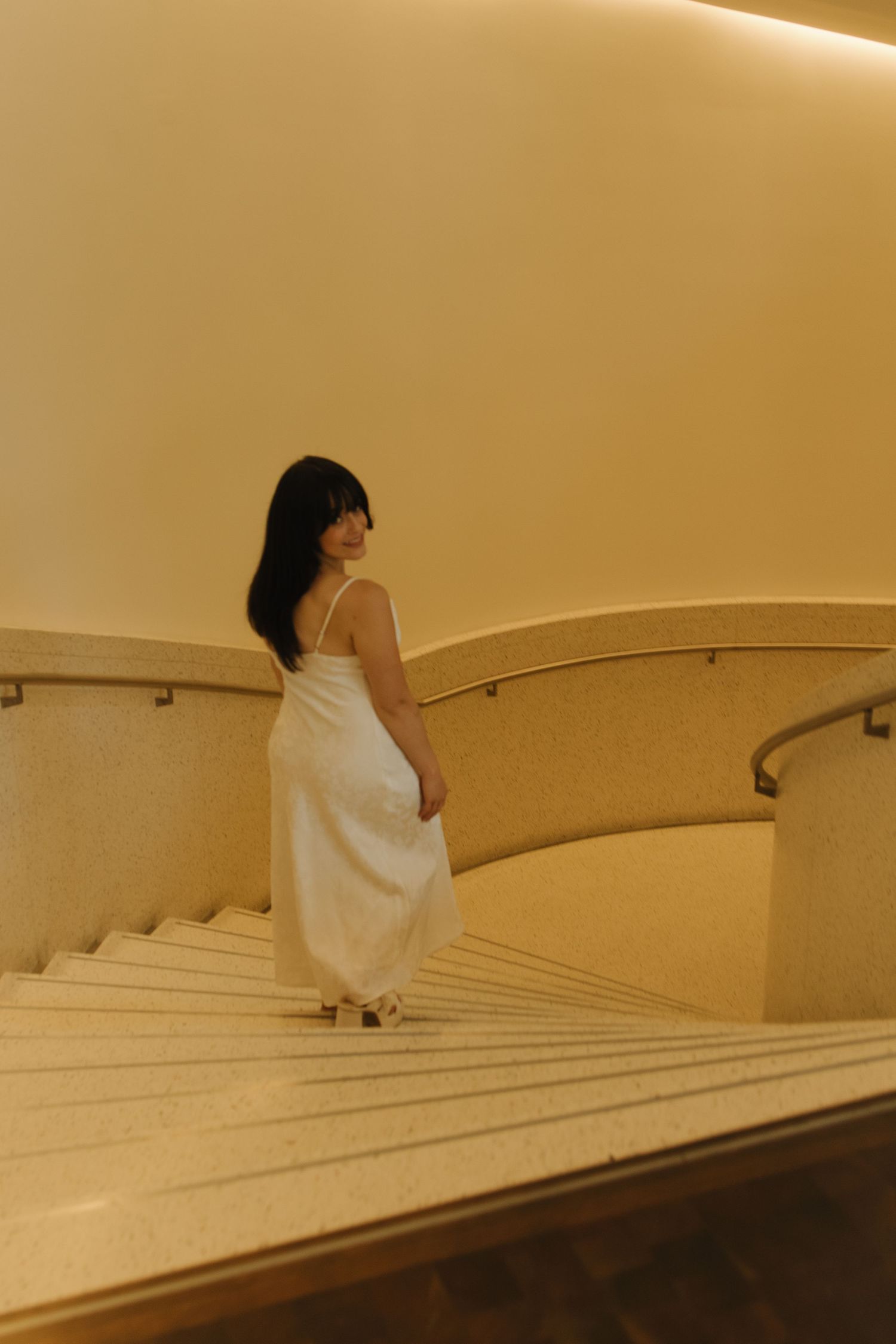 Person in white dress descending curved marble stairs in warm ambient lighting.