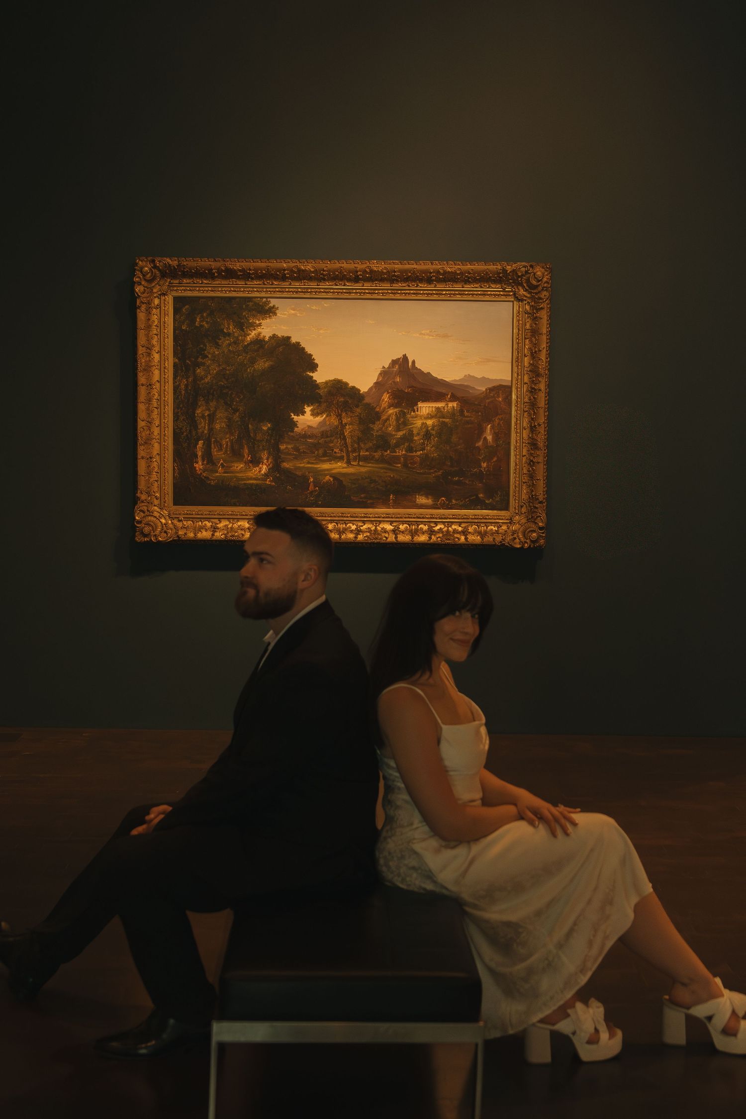 Couple in formal attire sitting and viewing artwork in a museum's dimly lit gallery space.