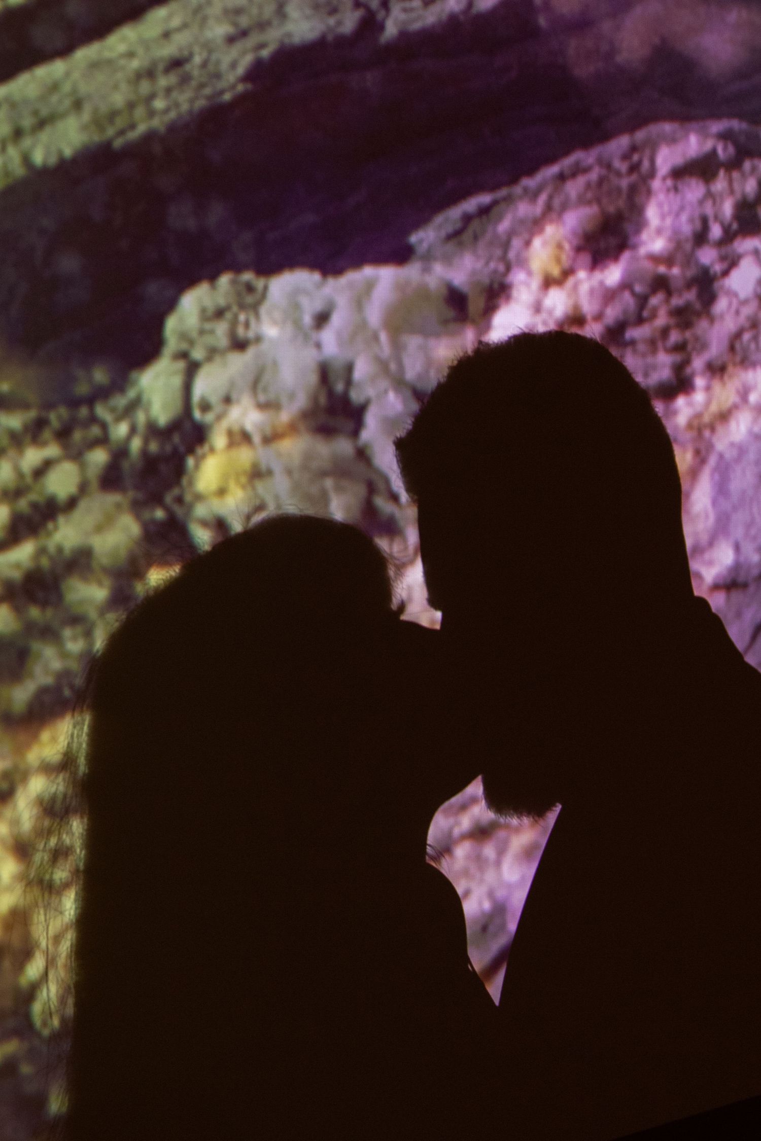 Silhouetted figures in an intimate moment against pink and purple lit rocky background.