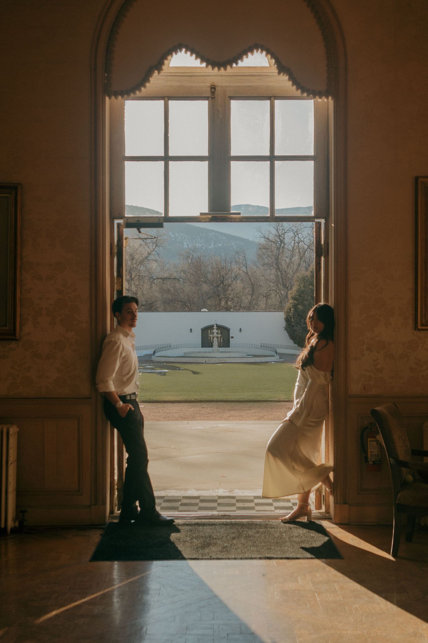 A romantic couple stands silhouetted in sunlight streaming through ornate windows of a grand historic building.