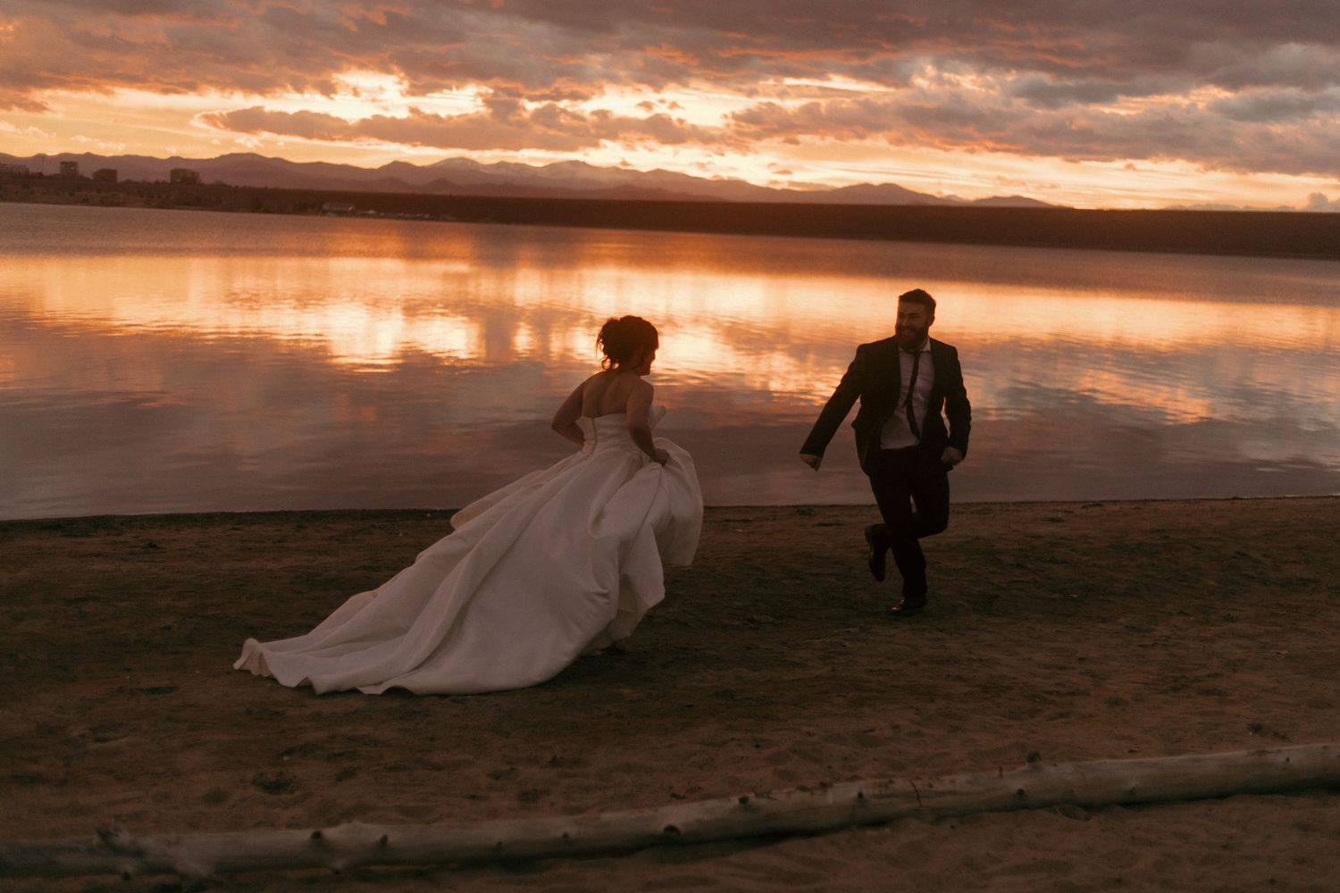 A couple in wedding attire walks along a lakeside at sunset with dramatic clouds reflecting on the water.