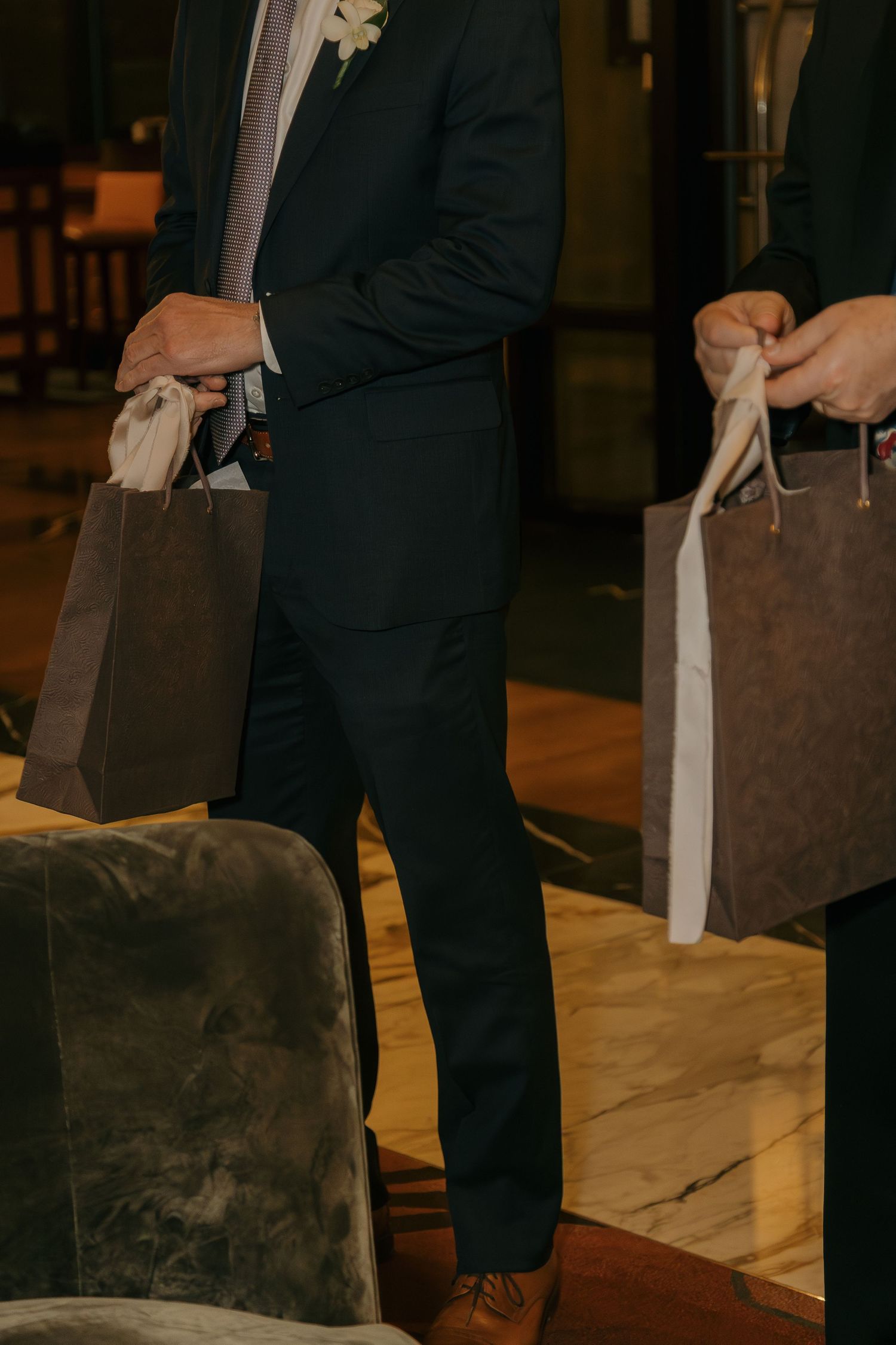 Man in dark suit holding brown gift bags at a formal event, wearing brown dress shoes and floral boutonniere.