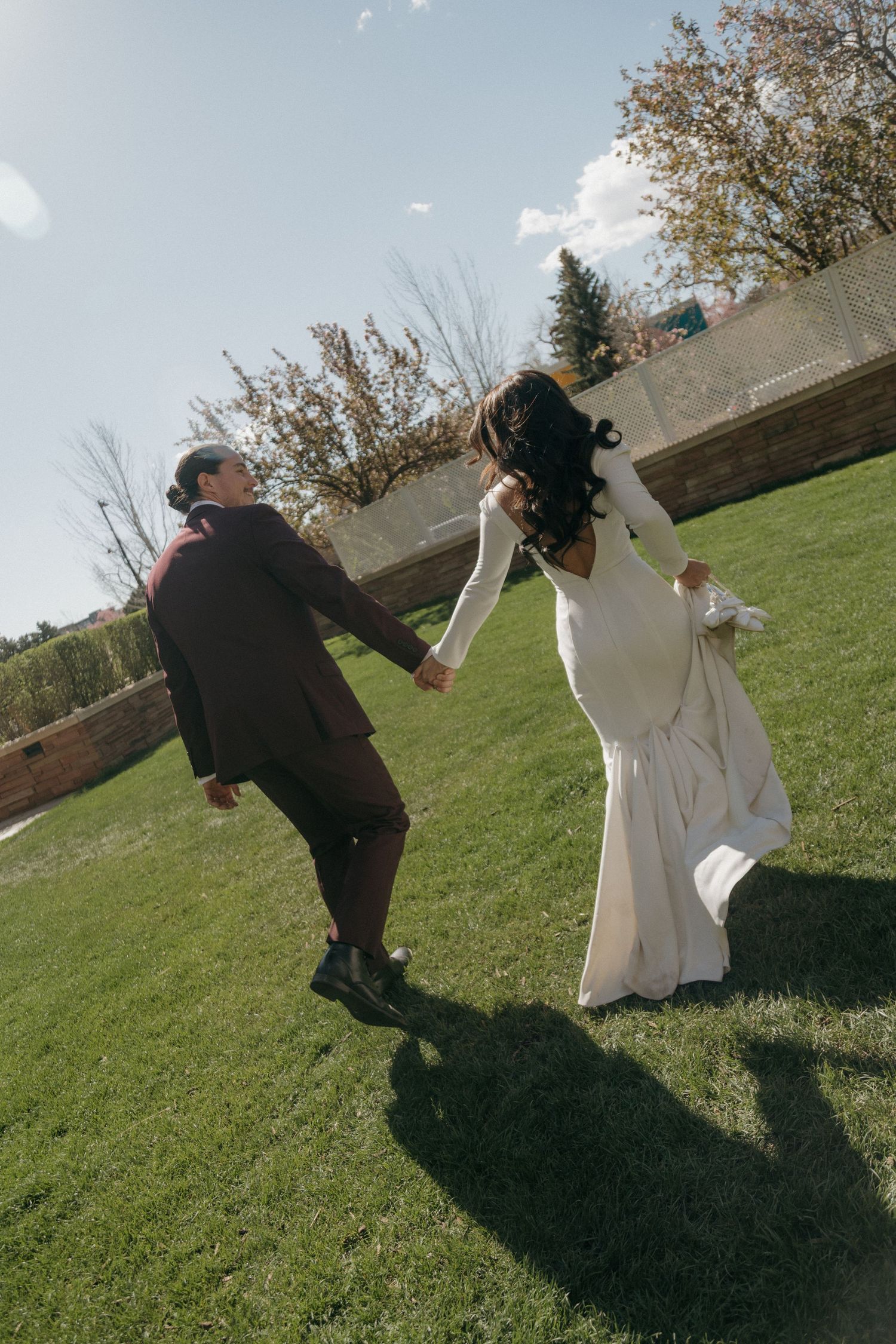 Bride in white mermaid gown and groom in burgundy suit hold hands dancing on green lawn under blue sky.