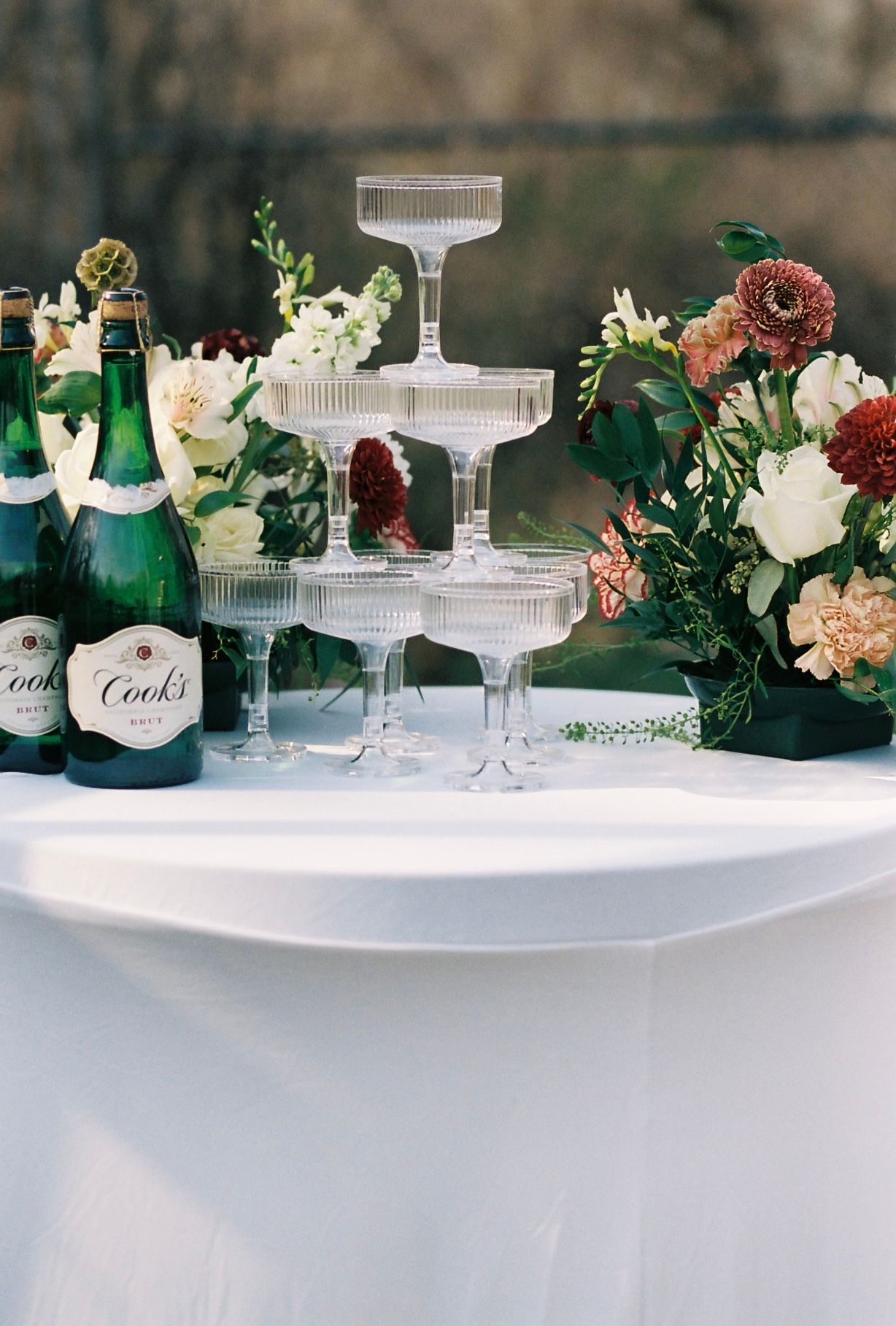 Champagne tower with Cook's bottles and lush floral arrangement on white tablecloth at wedding reception.