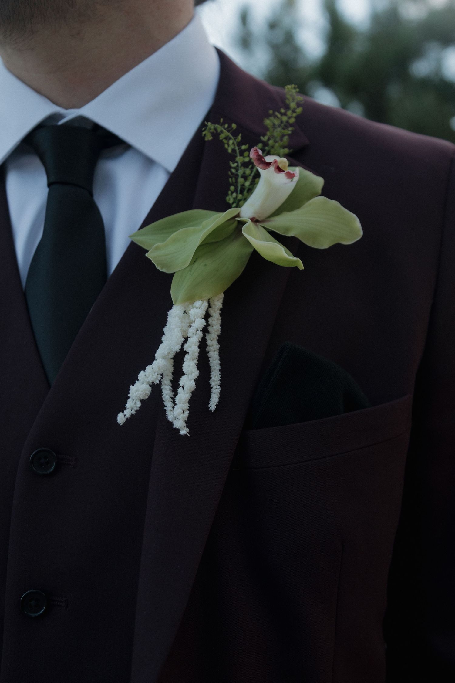 Close-up of a groom's burgundy suit boutonniere featuring green orchids and white hanging amaranthus blooms.
