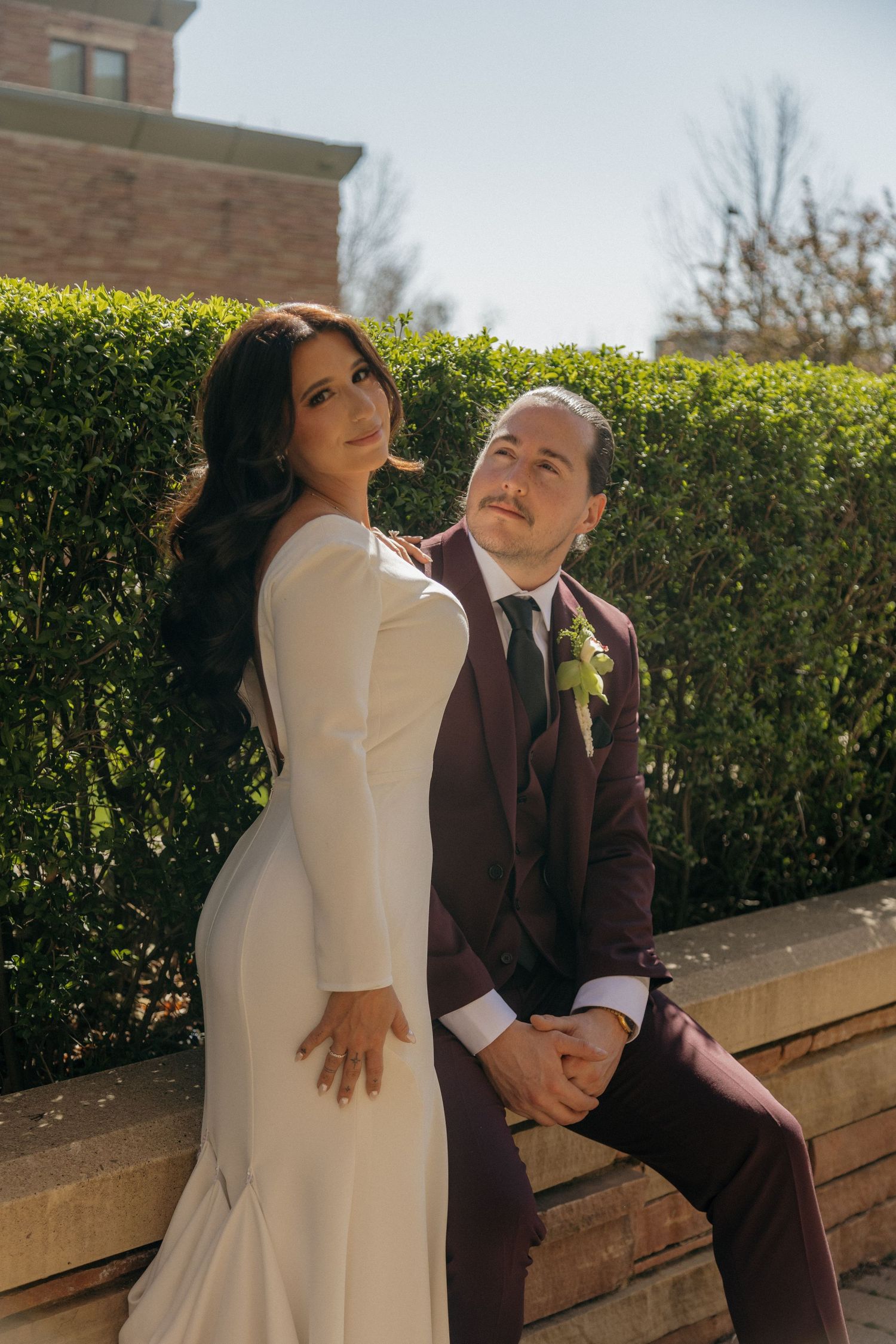Bride in white long-sleeve gown and groom in burgundy suit pose together outdoors near green hedges on their wedding day.