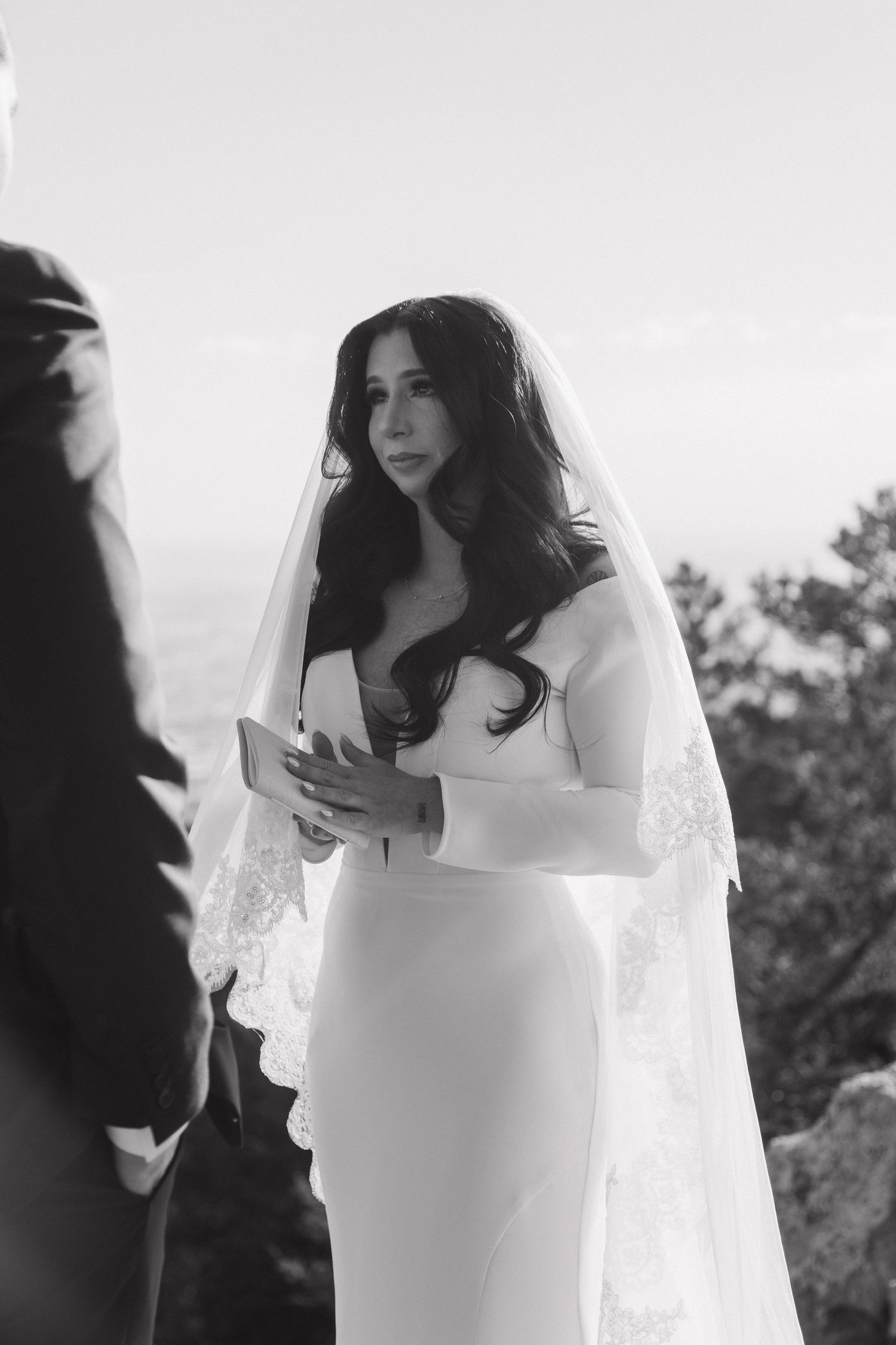 Bride in white strapless gown and lace veil reading vows during outdoor wedding ceremony.