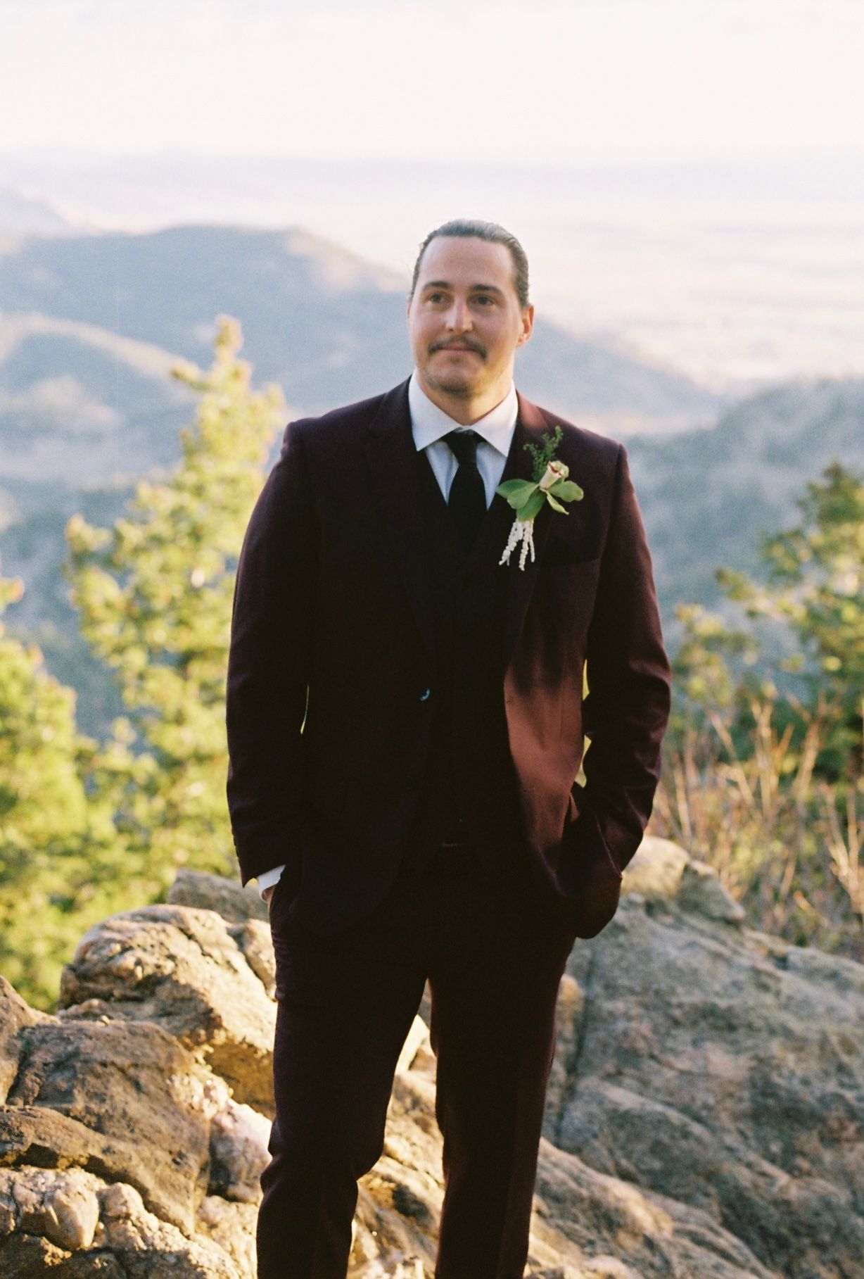Groom in burgundy three-piece suit with boutonniere stands on rocky mountain overlook at sunset.
