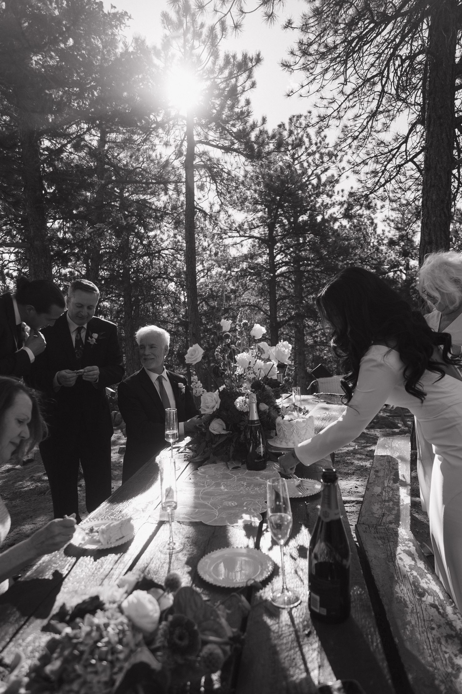 Black and white photo of wedding guests gathered around a rustic wooden table with floral arrangements in a forest setting.