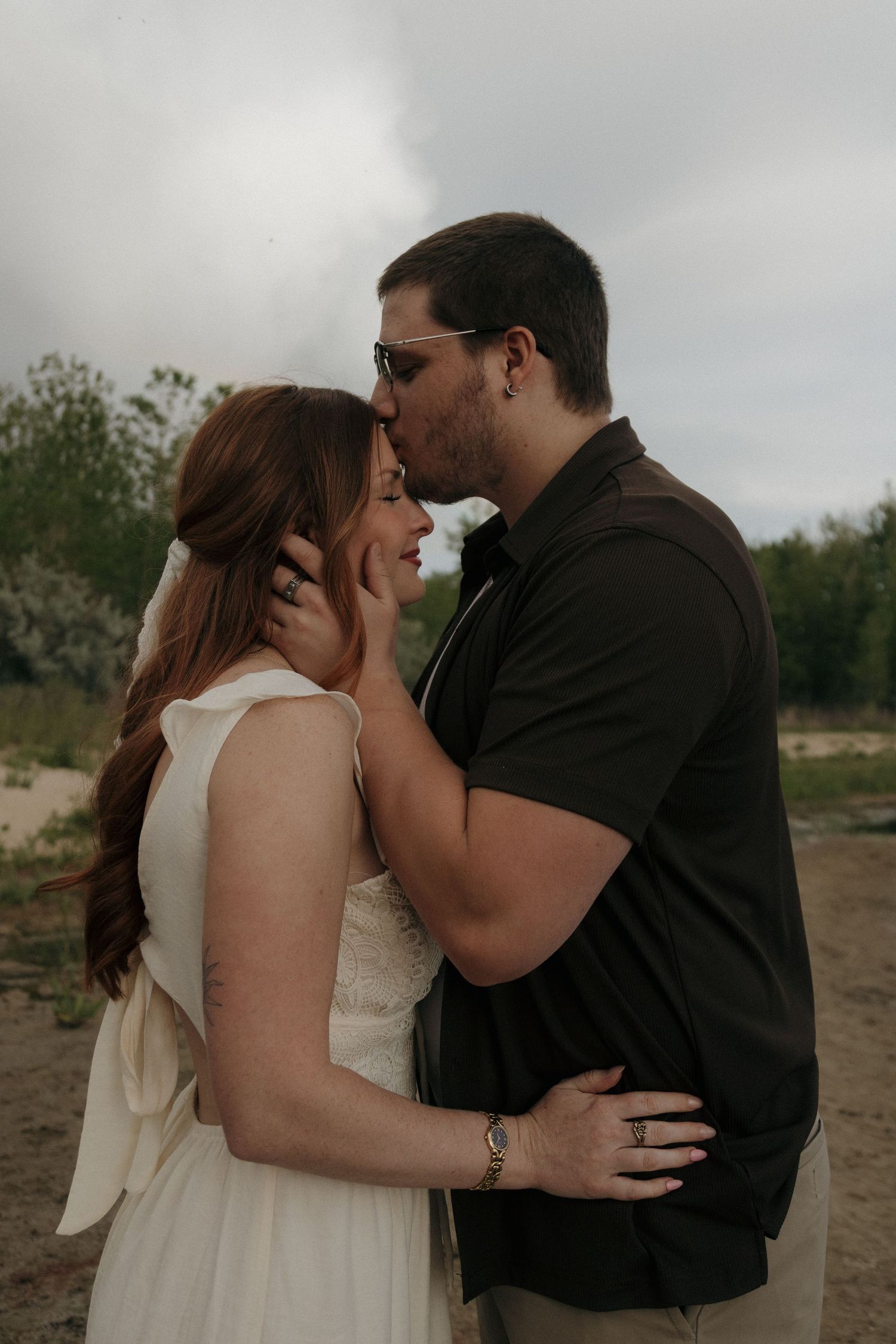 Couple sharing a romantic kiss outdoors, bride in white dress and groom in black shirt embracing on a sandy backdrop.