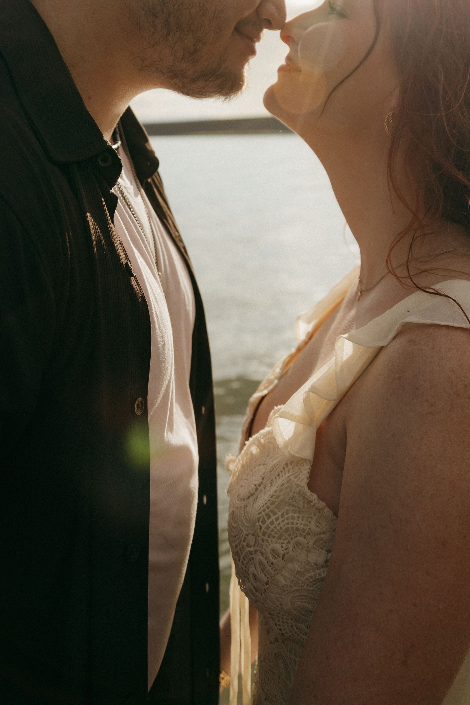 Couple sharing a tender almost-kiss in warm backlit sunlight near water.