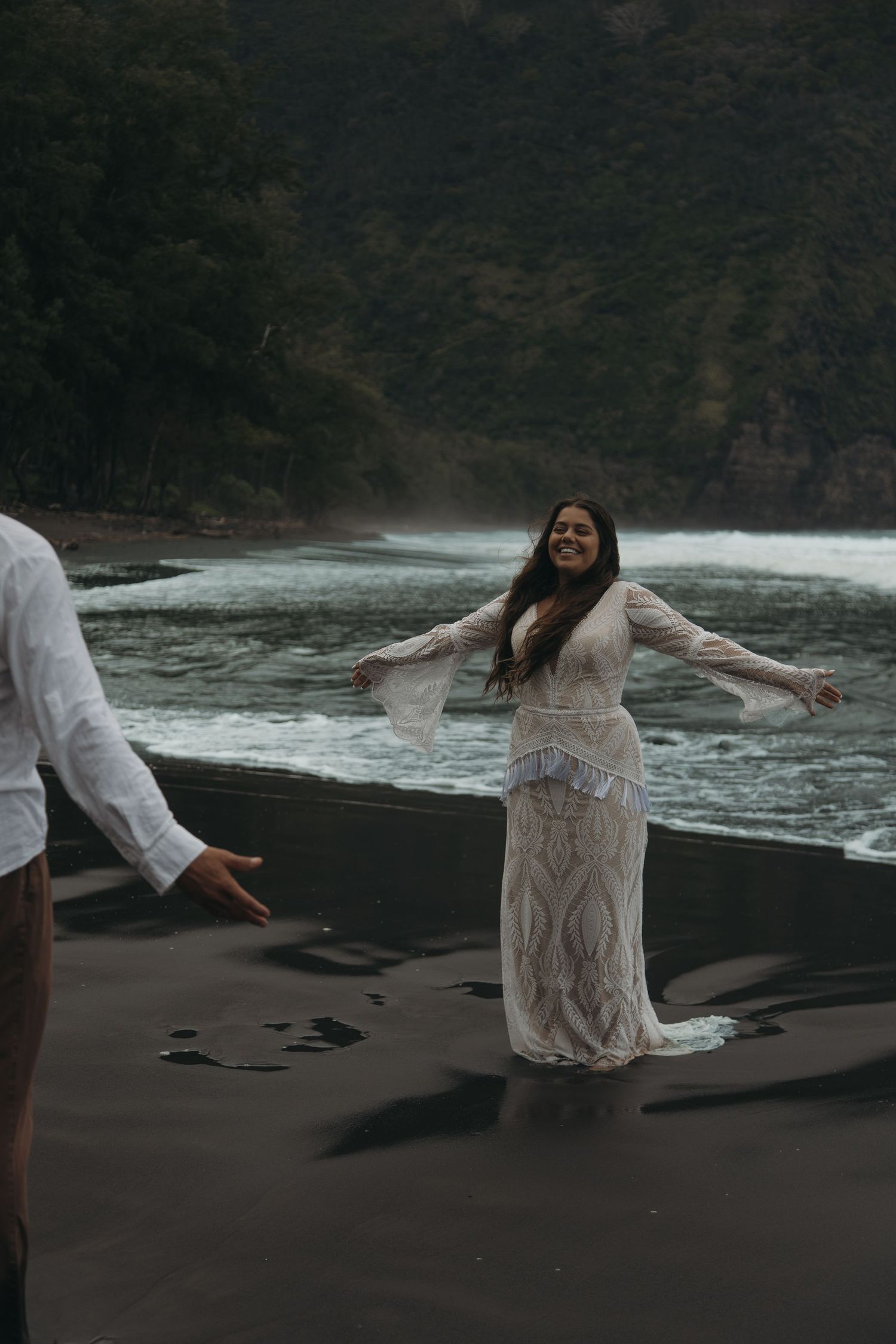 Joyful bride in white lace dress twirls on black sand beach with lush green cliffs and river behind her.