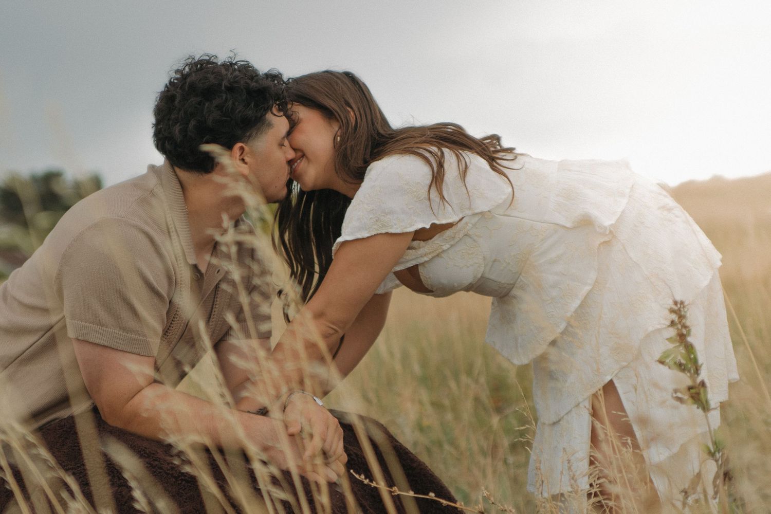 Couple leans in for a kiss surrounded by tall golden grasses in warm sunlight.