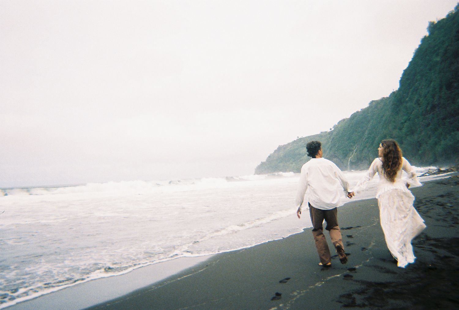 Bride and groom holding hands running along black sand beach with crashing waves and green cliffs.