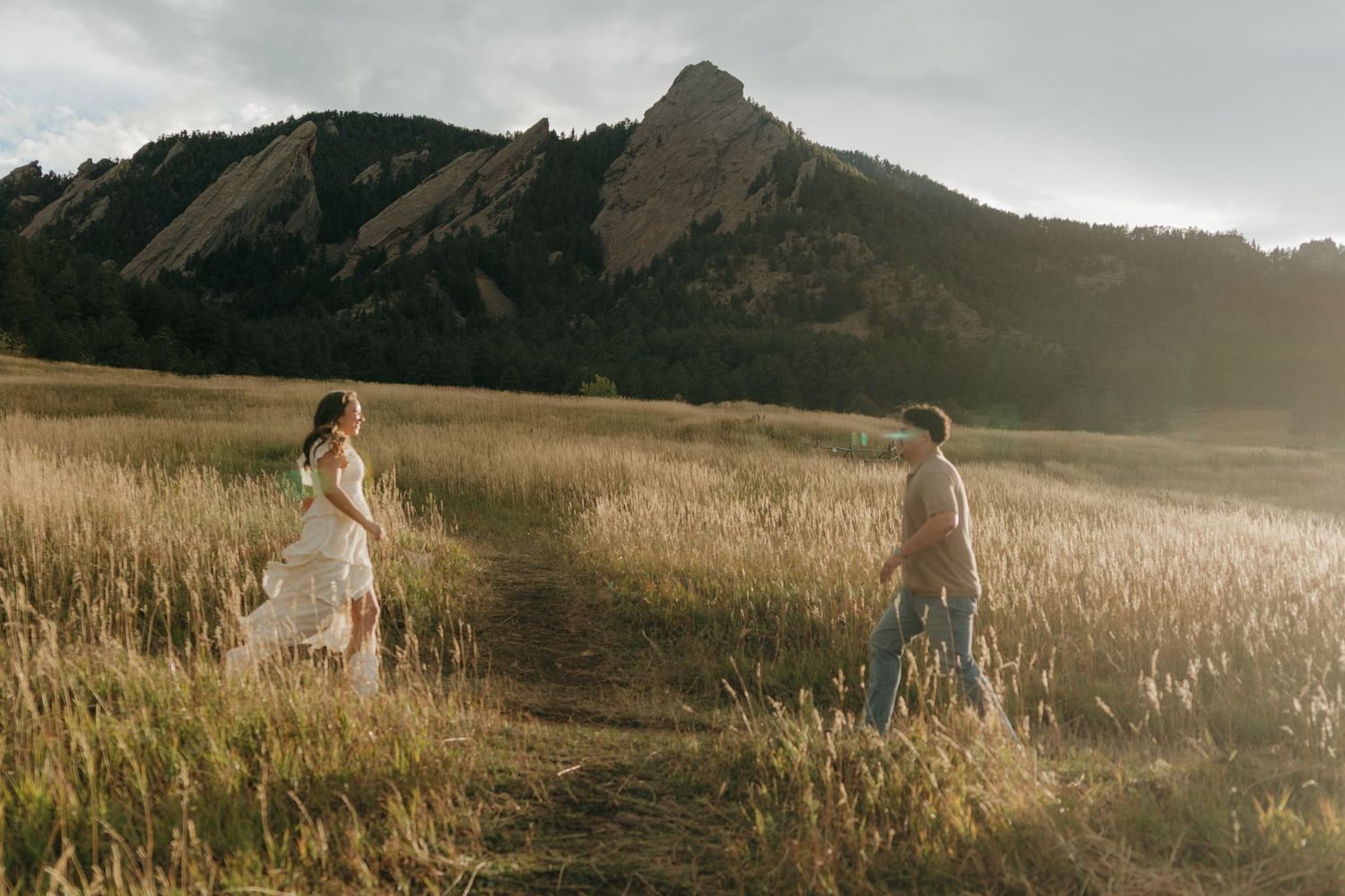 Couple walking through golden grass meadow at sunset with dramatic rocky mountain flatirons behind them.