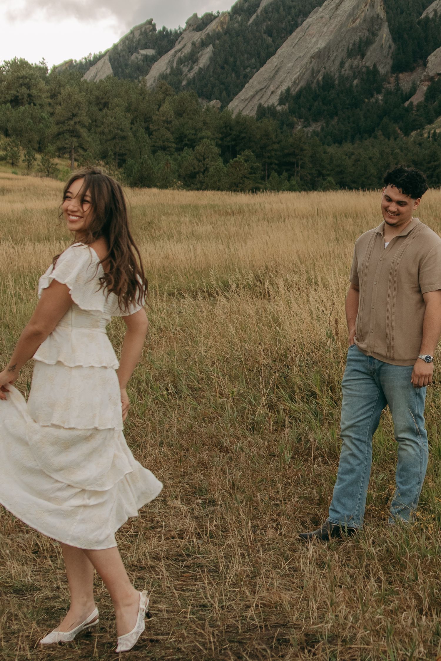 Joyful woman in white tiered dress twirling in a field while her partner smiles at the Flatirons.