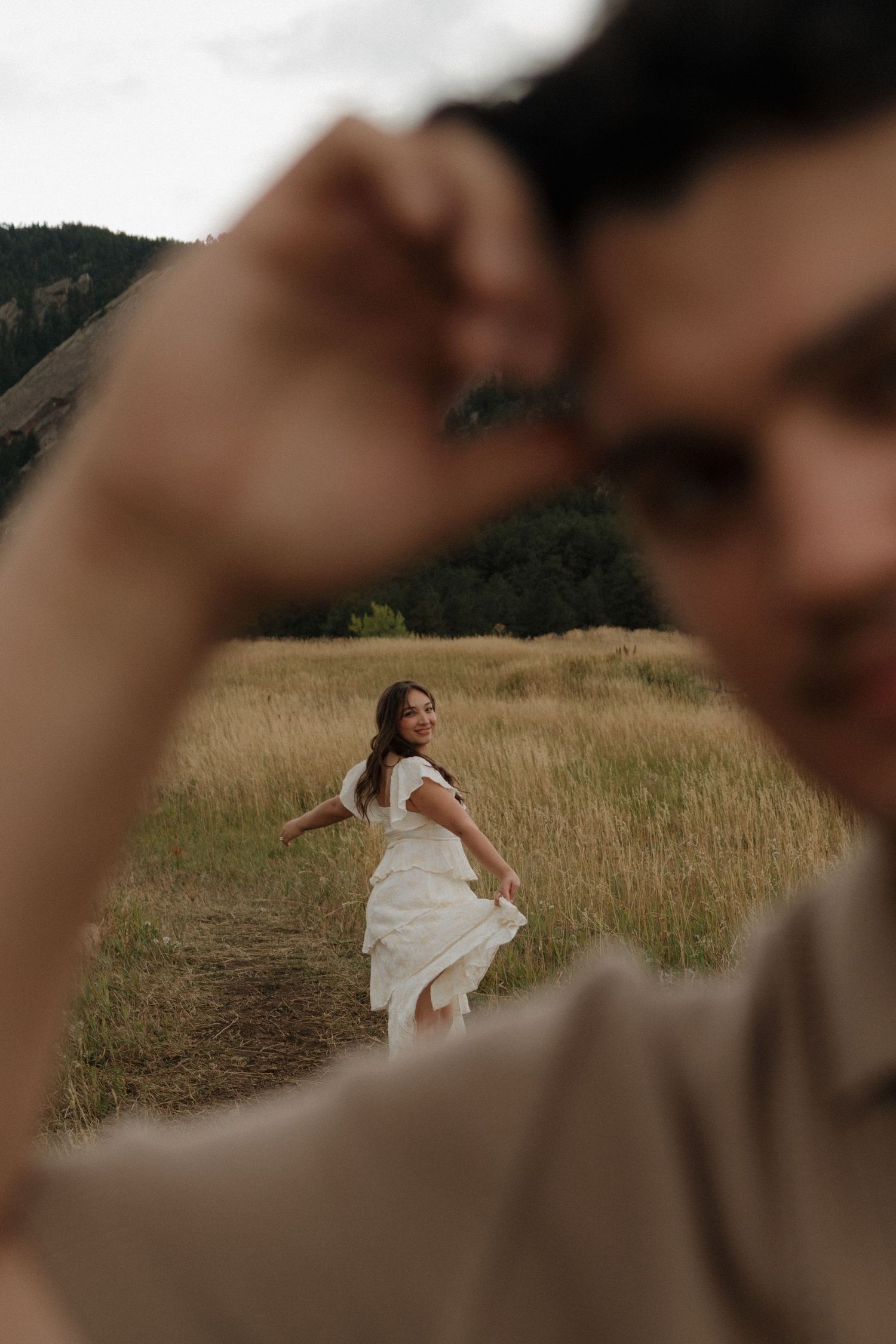 Bride in white tiered dress smiles while groom leads her through golden meadow near Colorado Flatirons.