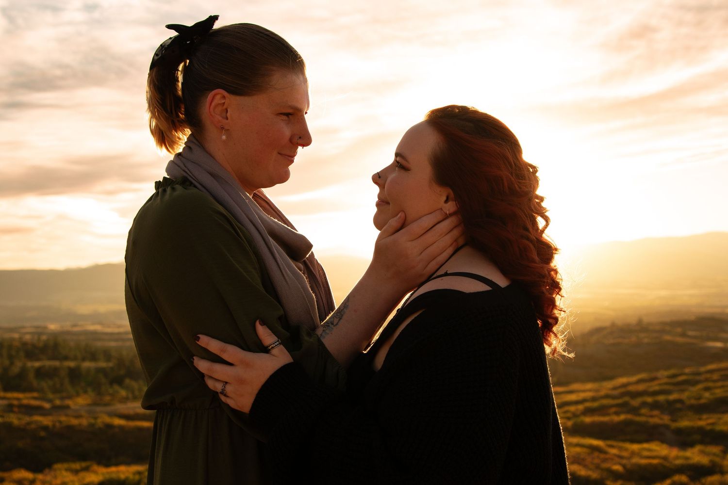 Two women share an intimate moment at golden hour, one tenderly holding the other's face against a scenic landscape.