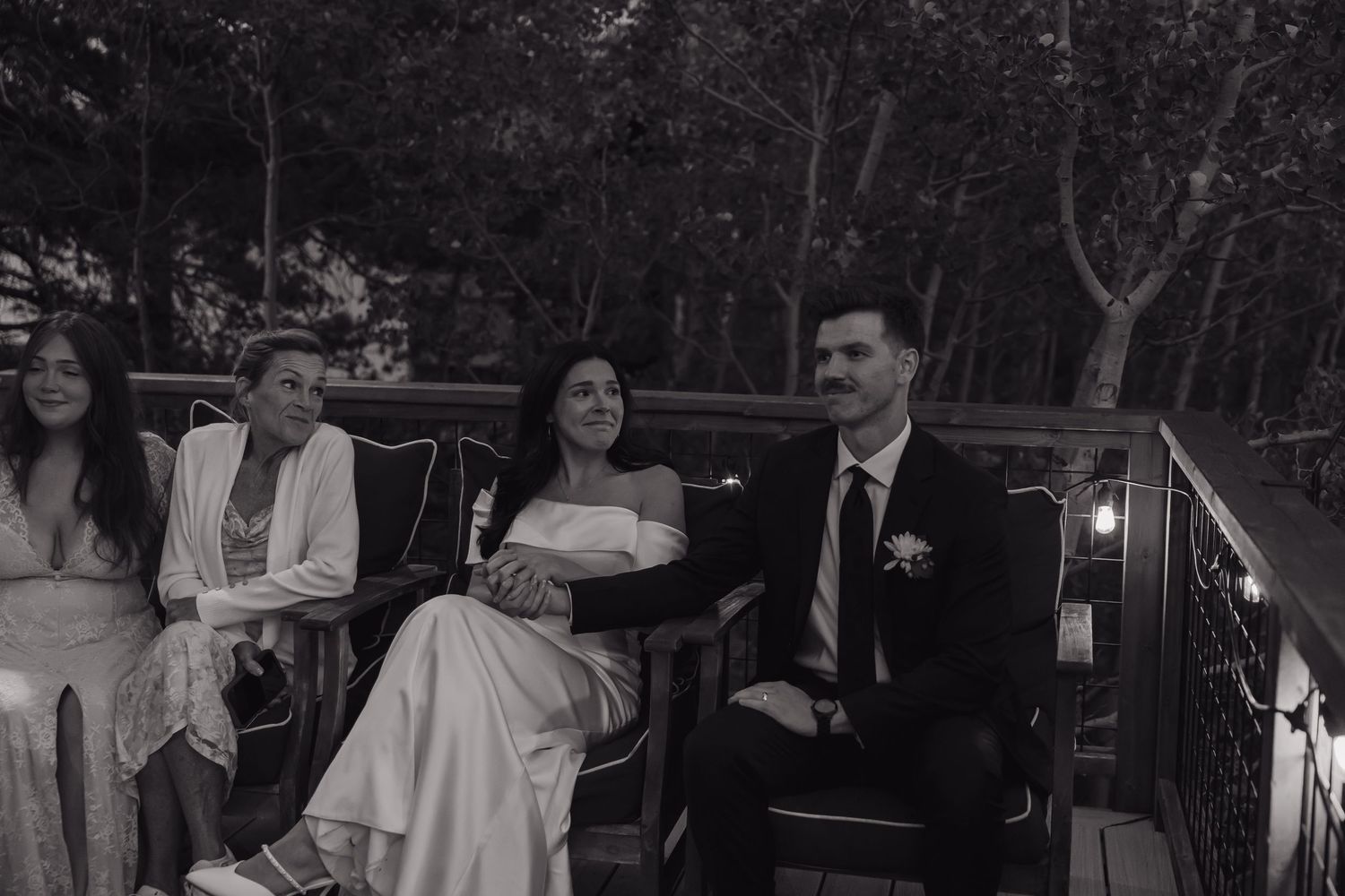 Black and white photo of bride and groom seated with guests on outdoor deck at wedding.