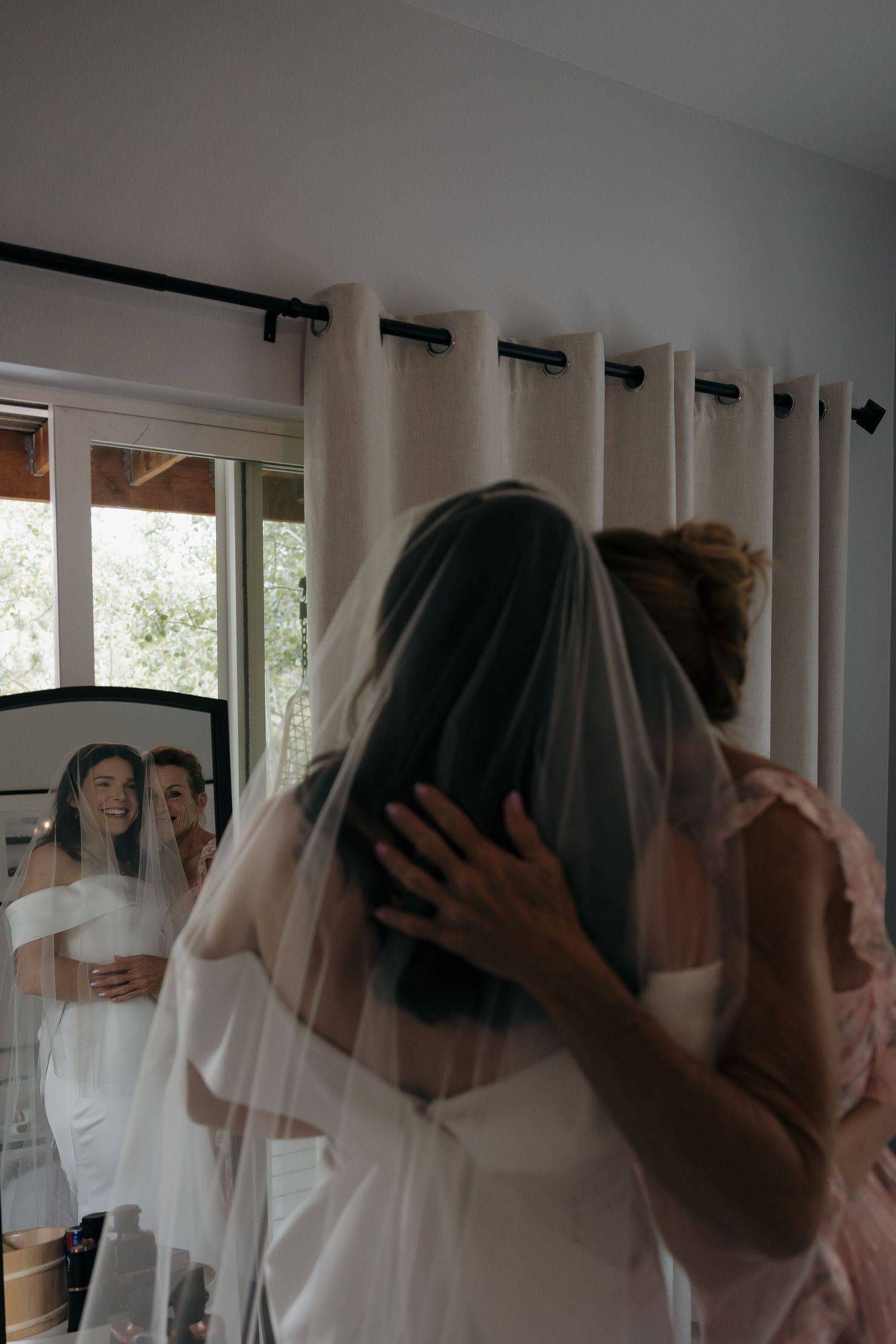 Bride in white gown and veil being helped by bridesmaid, with mirror reflection showing her smiling face.