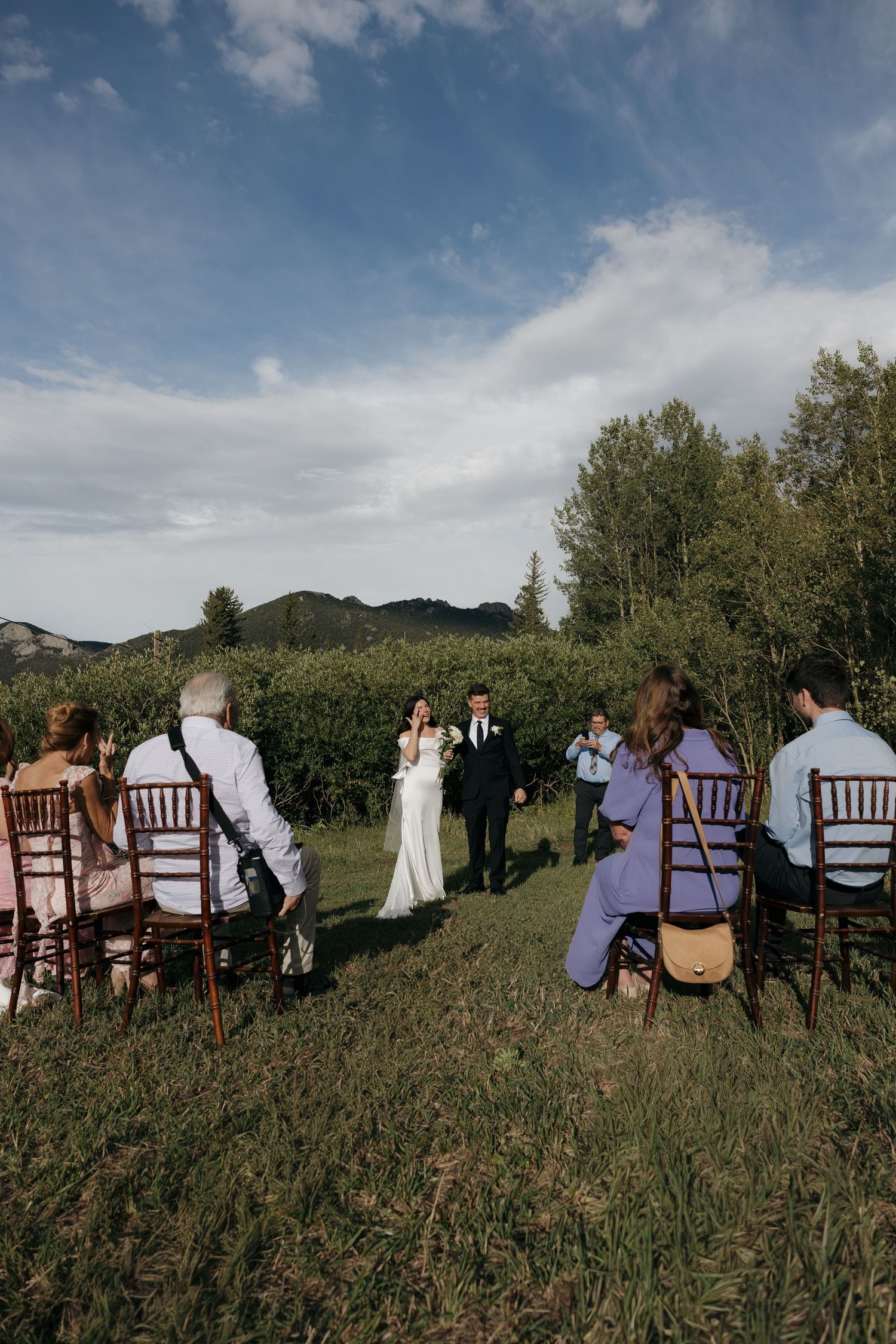 Intimate outdoor wedding ceremony in a mountain meadow with guests seated on wooden chairs facing the couple.