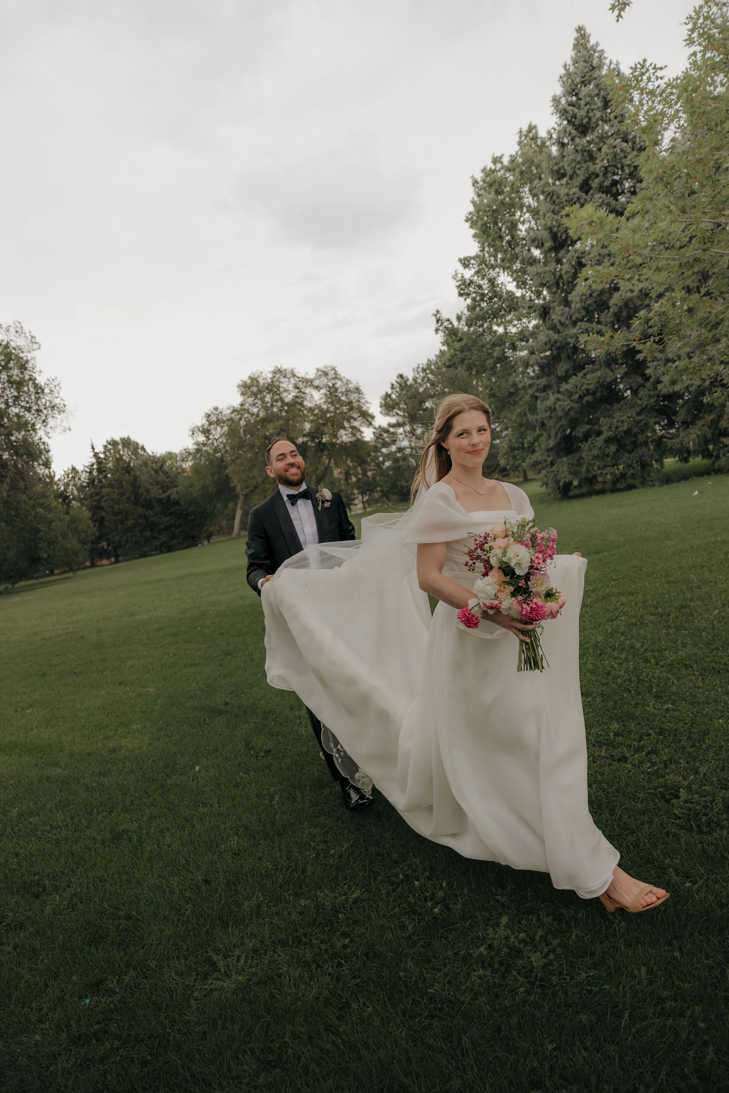 Bride in flowing white gown holding pink bouquet walks with groom in tuxedo on green lawn.