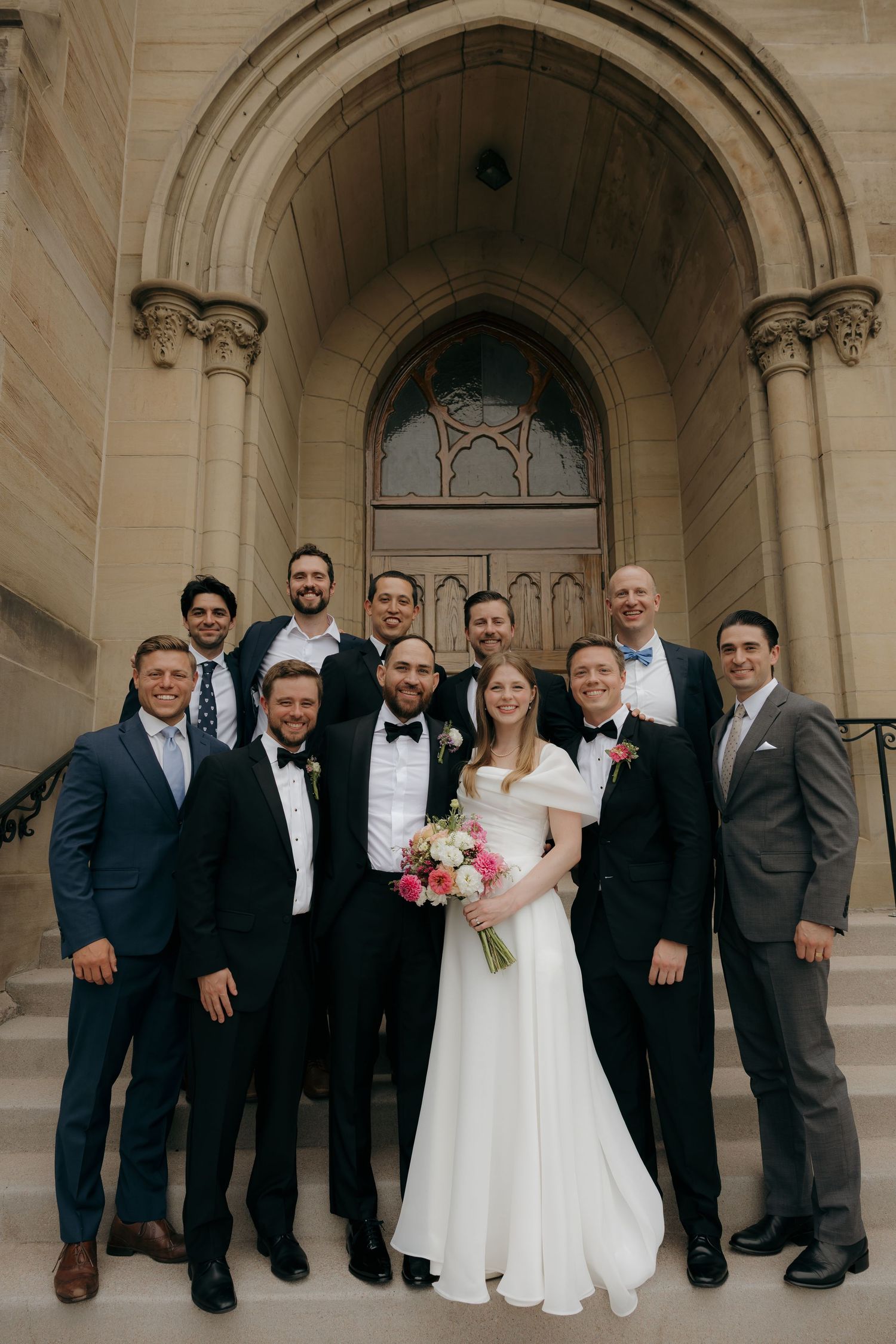 Wedding party poses on church steps with bride in white gown holding pink bouquet surrounded by groomsmen in tuxedos.