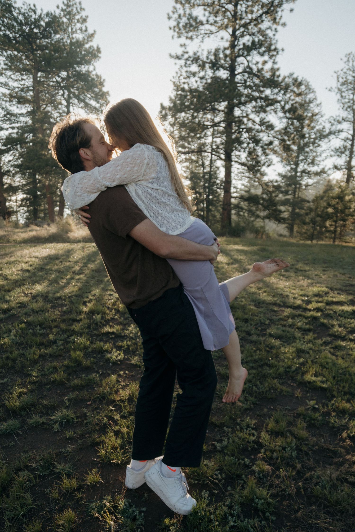 Man lifting and kissing woman in a sunny pine forest during a romantic outdoor engagement session.