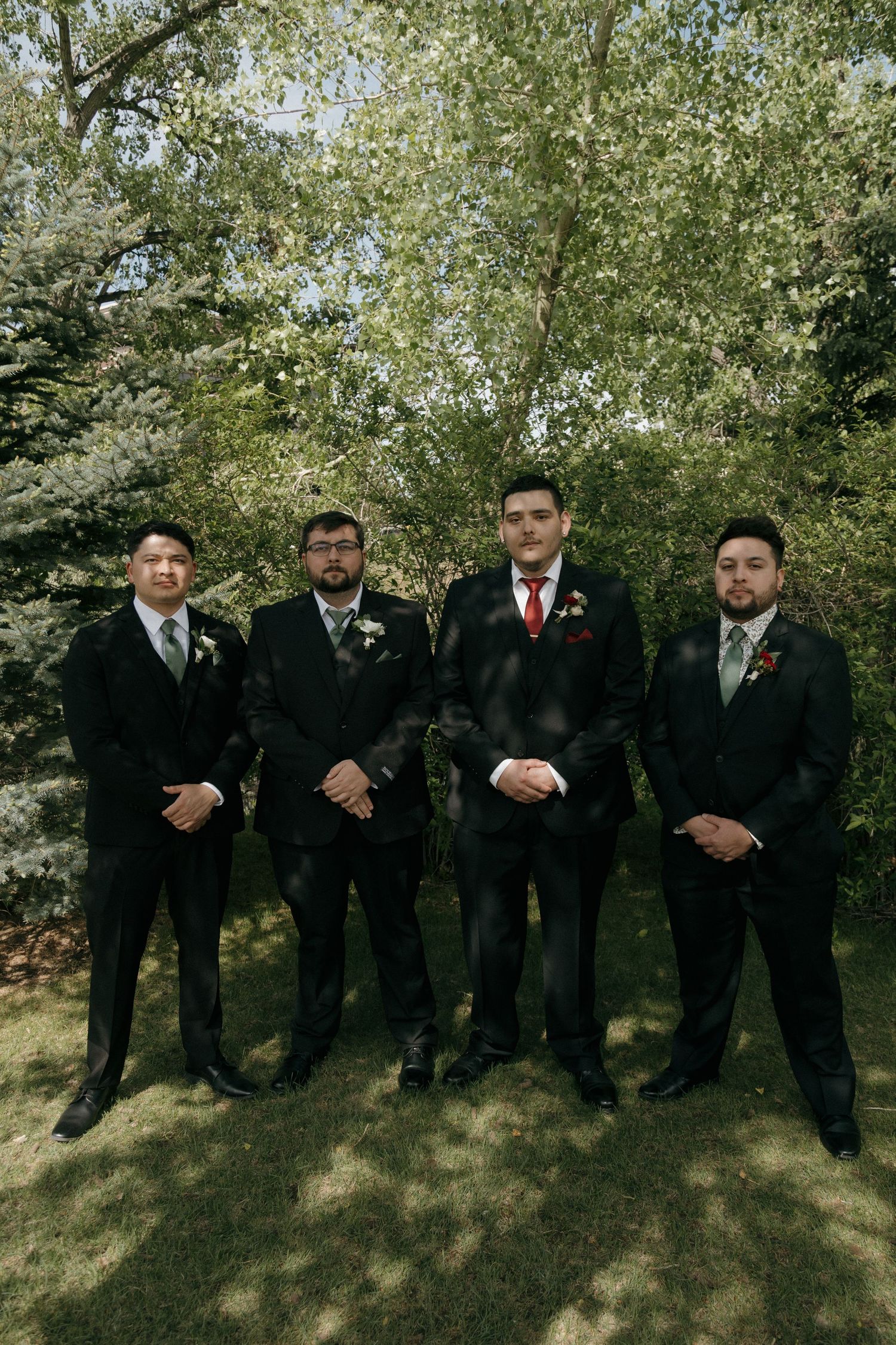 Four groomsmen in black suits with boutonnieres pose outdoors in front of lush green trees at a wedding.