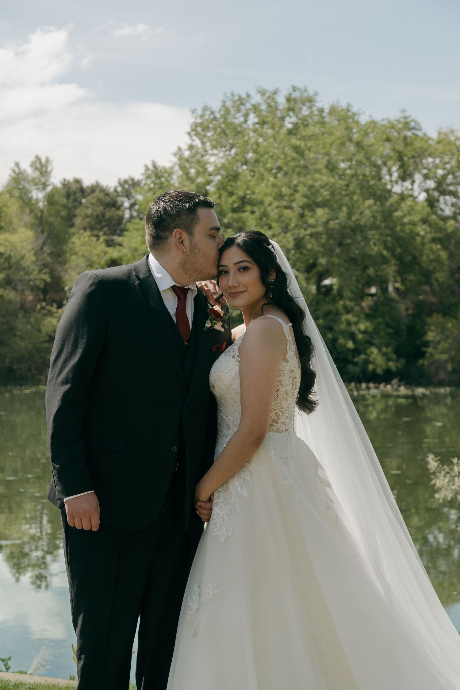 Groom kissing bride on cheek near a reflective pond with green trees.