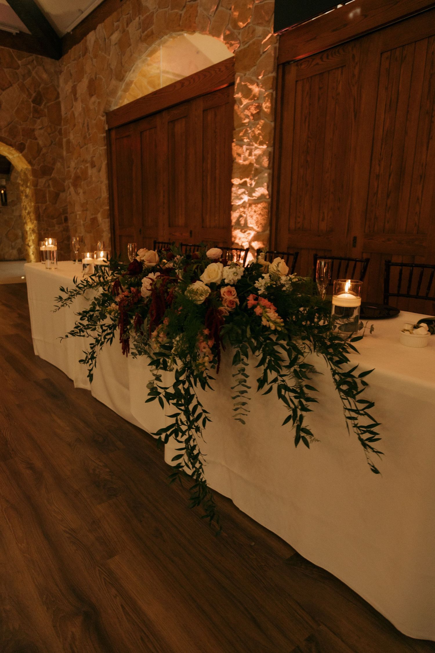 Romantic candlelit wedding head table with cascading floral centerpiece of roses and greenery in a rustic stone venue.