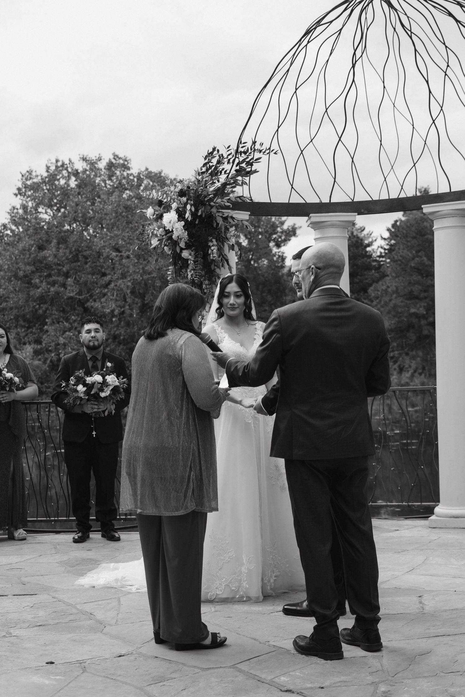Black and white wedding ceremony with bride, officiant, and groom exchanging vows under decorative arch outdoors.