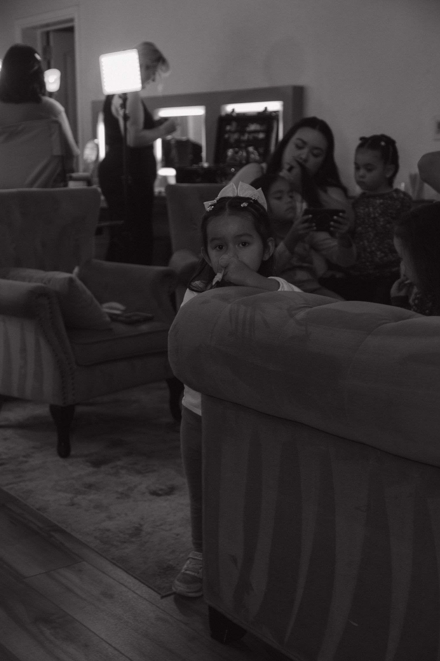 Black and white photo of young flower girl peeking over sofa arm while bridal party prepares in background.