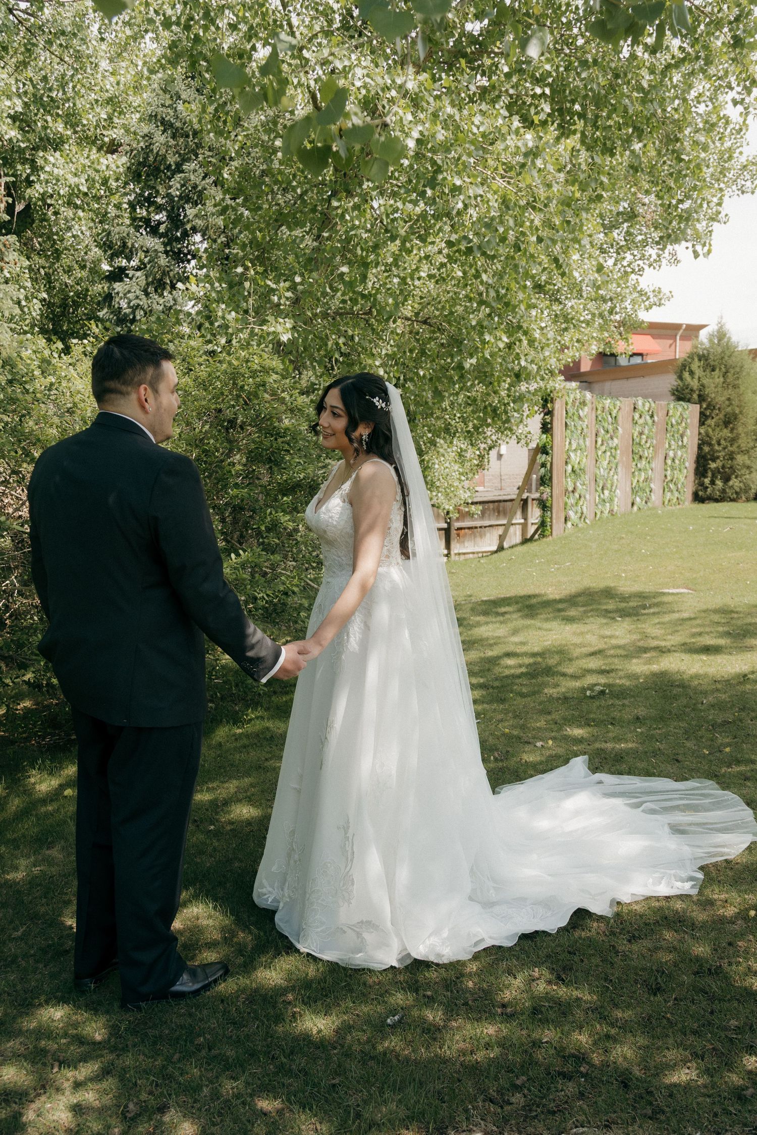Bride and groom hold hands in a lush green garden, bride wearing a flowing white gown with long train and veil.
