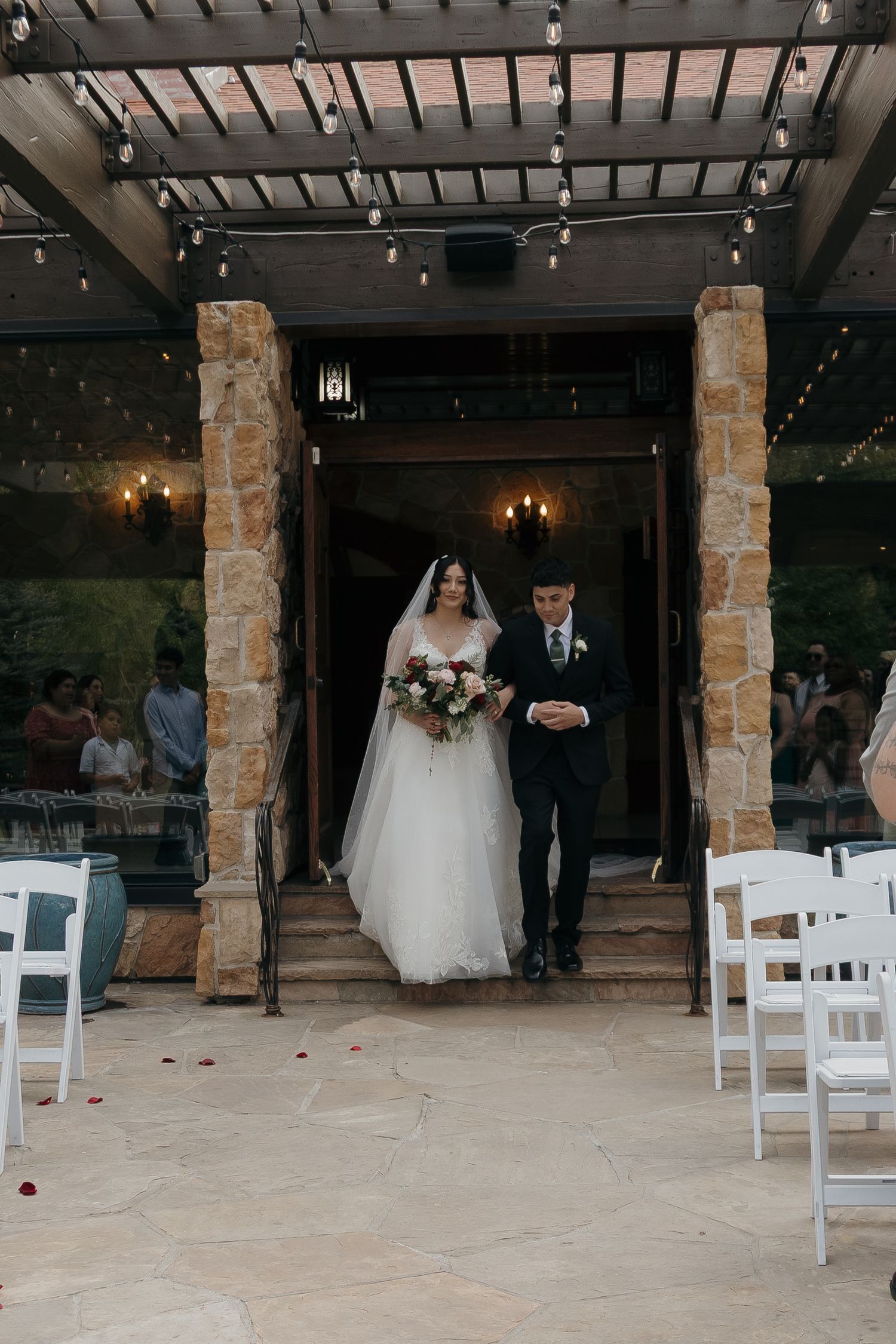 Bride holding bouquet walks down the aisle with escort through stone columns at an outdoor wedding venue.