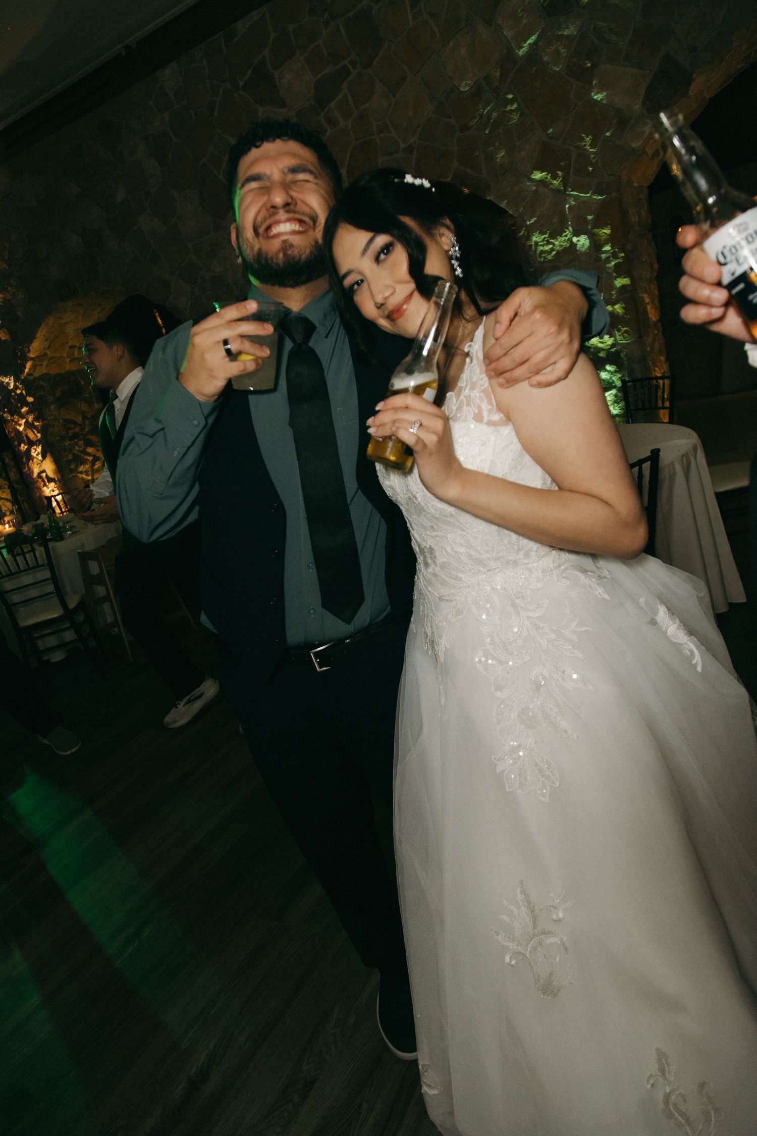 Bride in white lace gown and groom smiling and holding drinks together at wedding reception.
