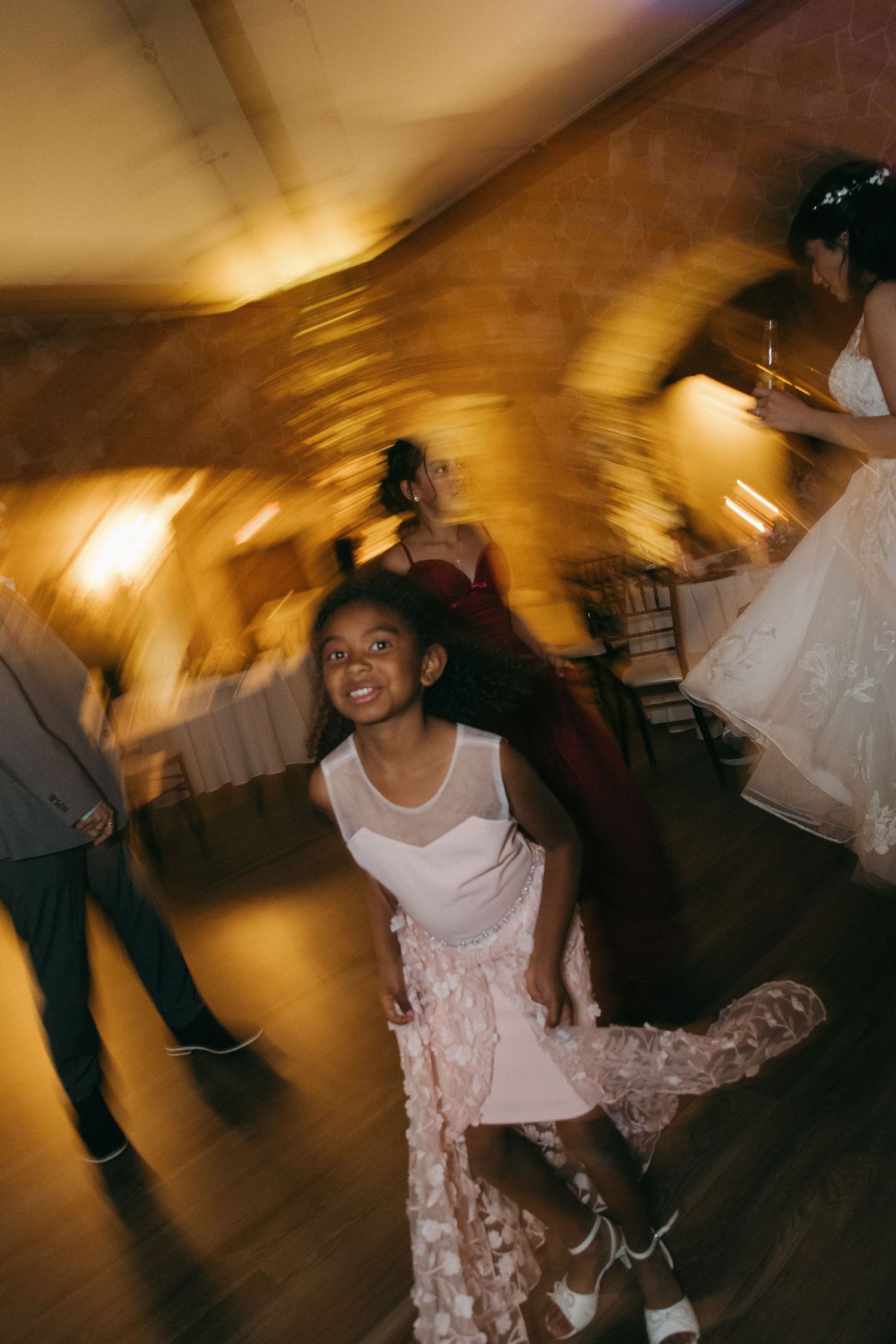 Young girl in pink dress twirling joyfully on a wedding reception dance floor with warm golden lighting.