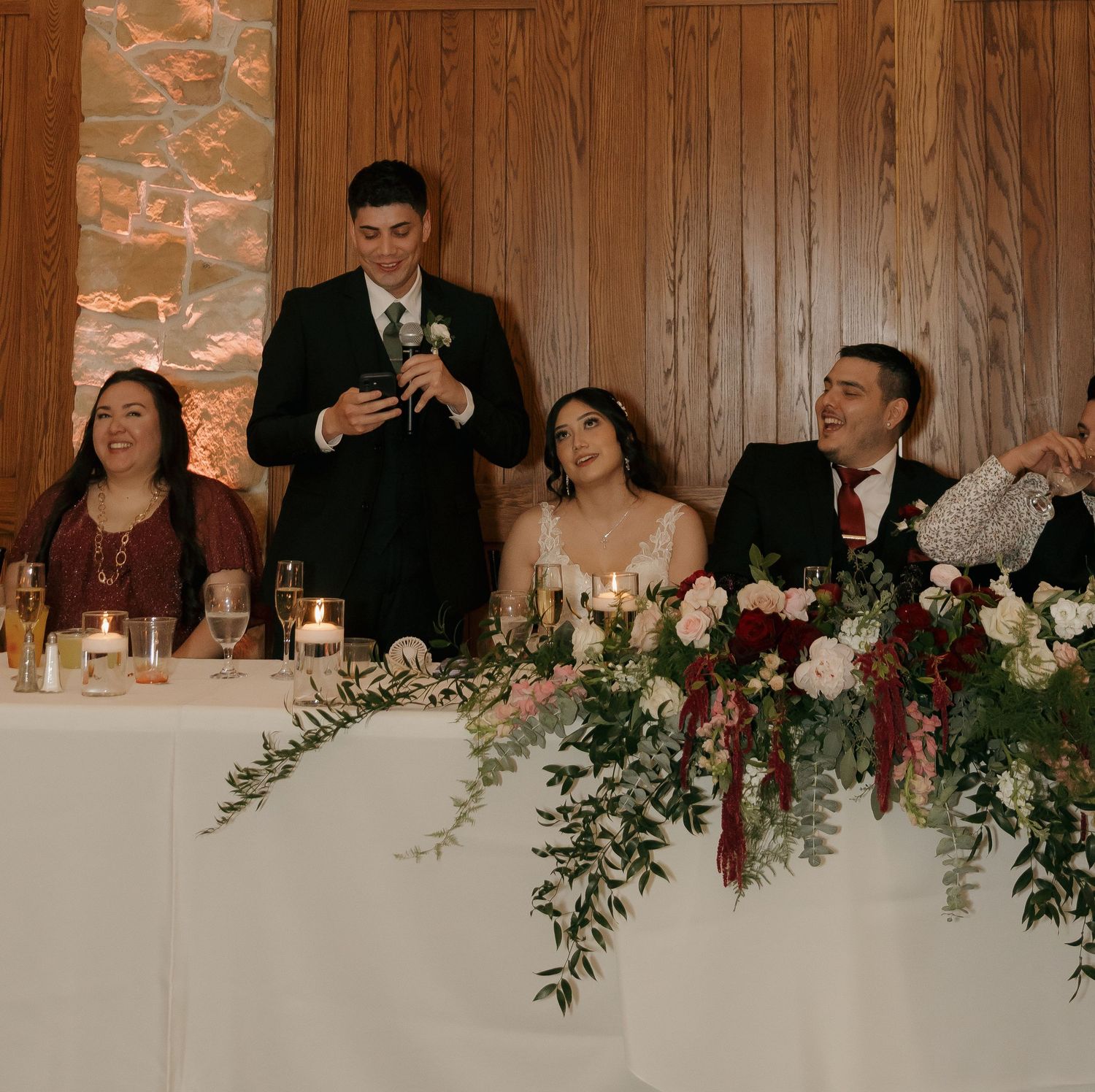 Wedding party laughs during heartfelt toast at elegant reception with floral centerpieces and candlelight.