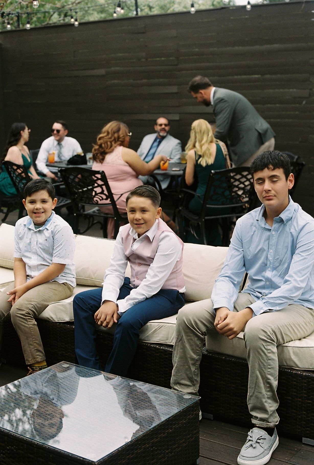 Three young boys in dress shirts sitting on patio furniture at an outdoor wedding reception.