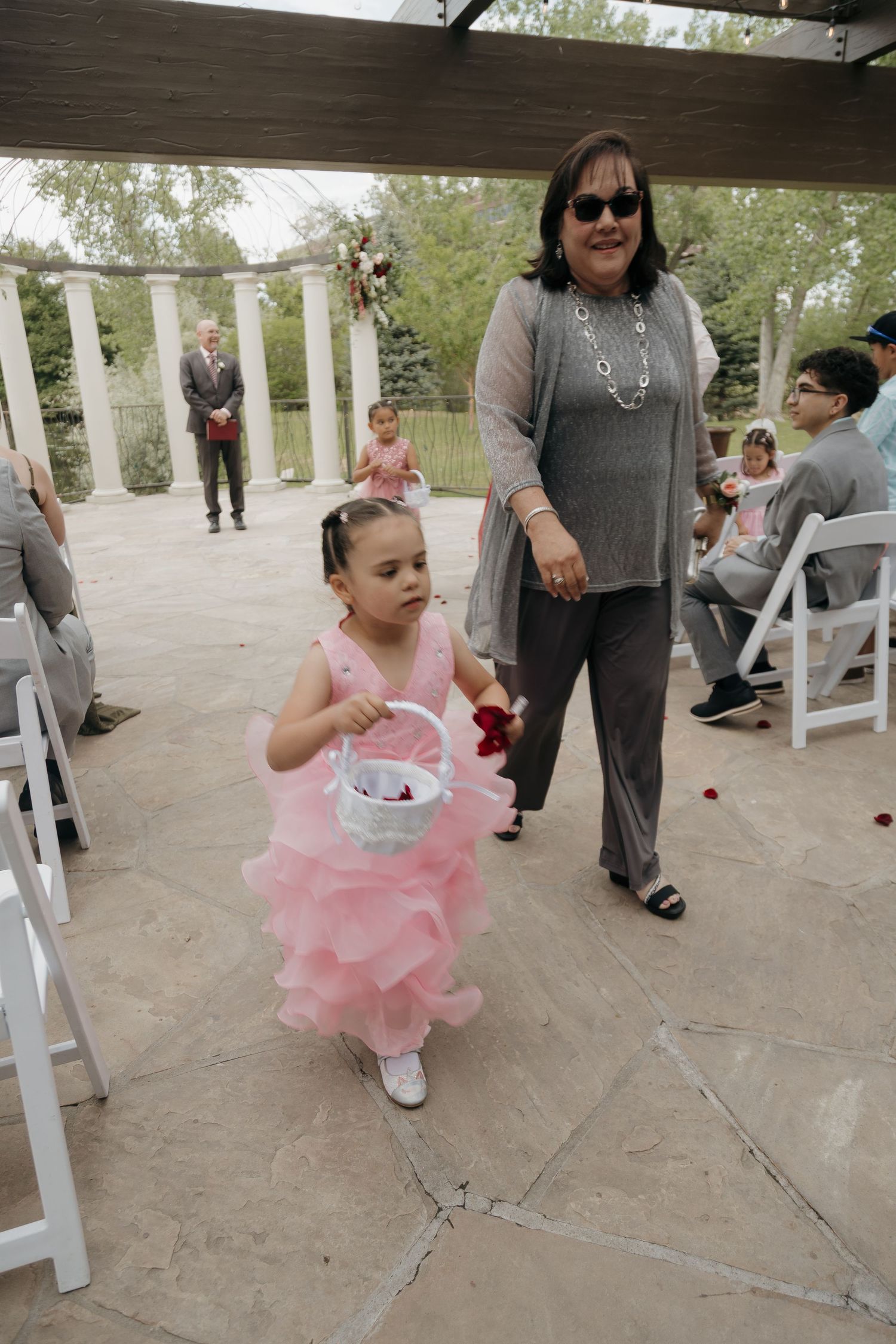 A flower girl in a pink dress carries a basket down the aisle at an outdoor wedding ceremony.