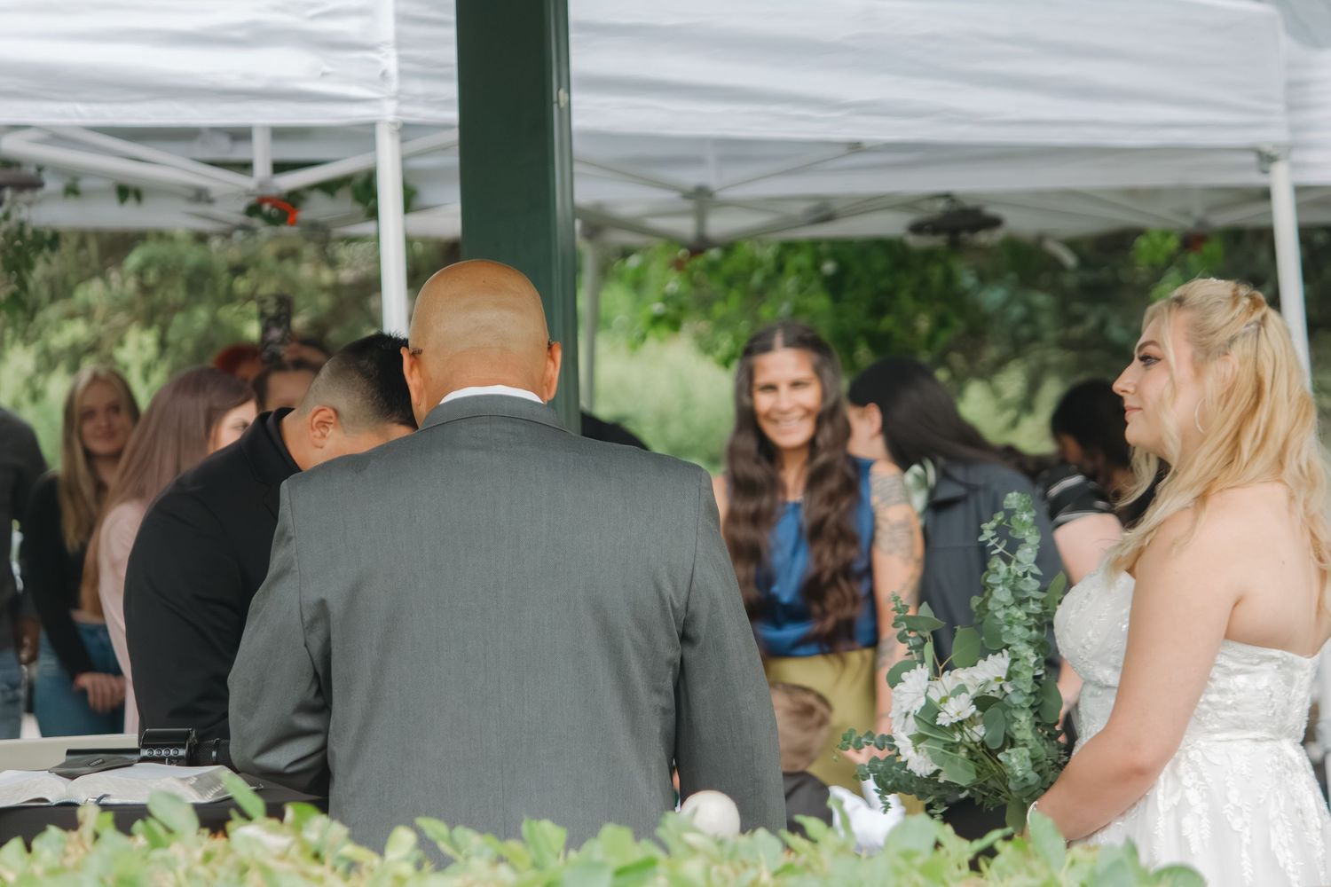 Wedding guests gather under a white tent during an outdoor reception.