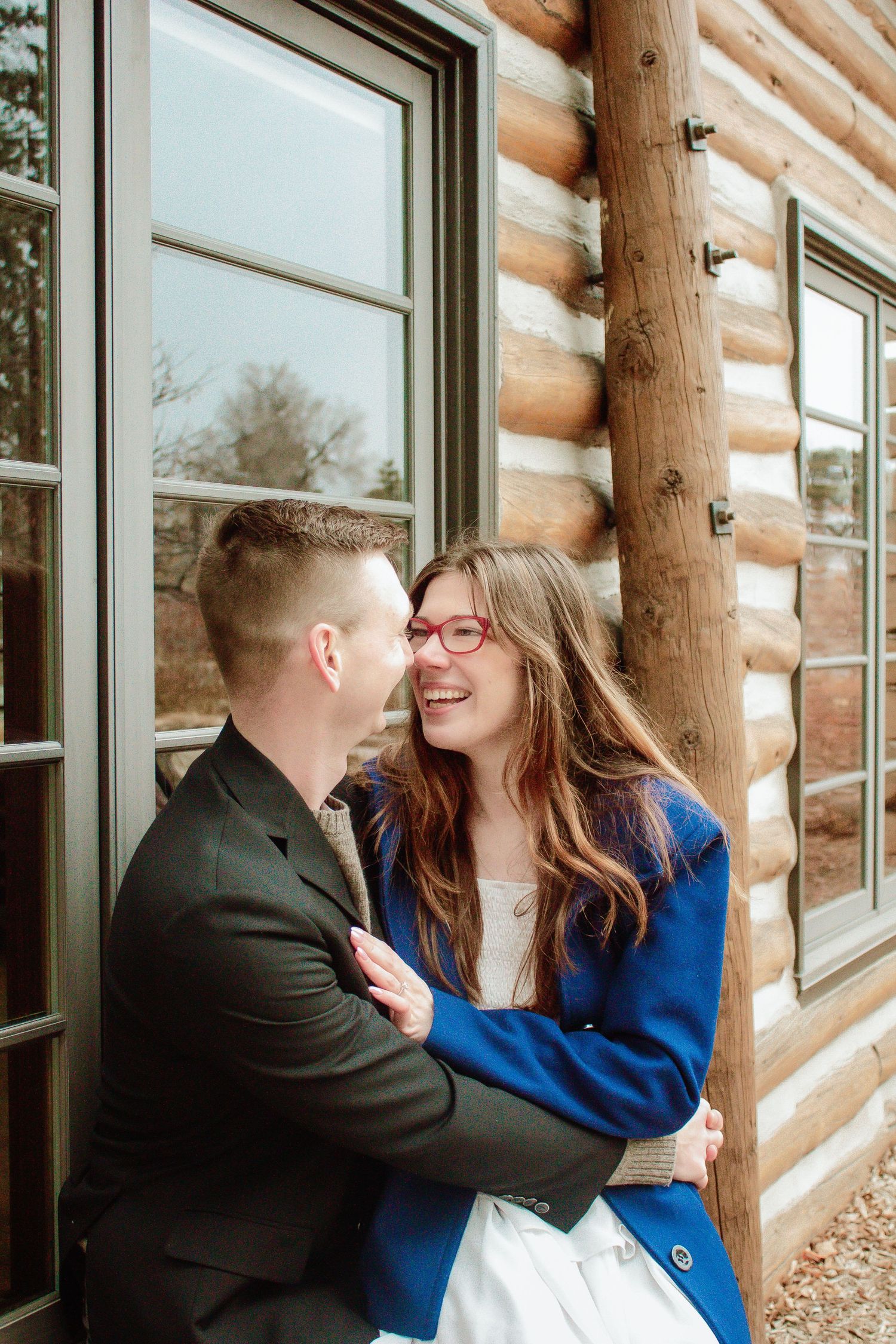 A couple shares a tender moment while leaning against a rustic log cabin wall.