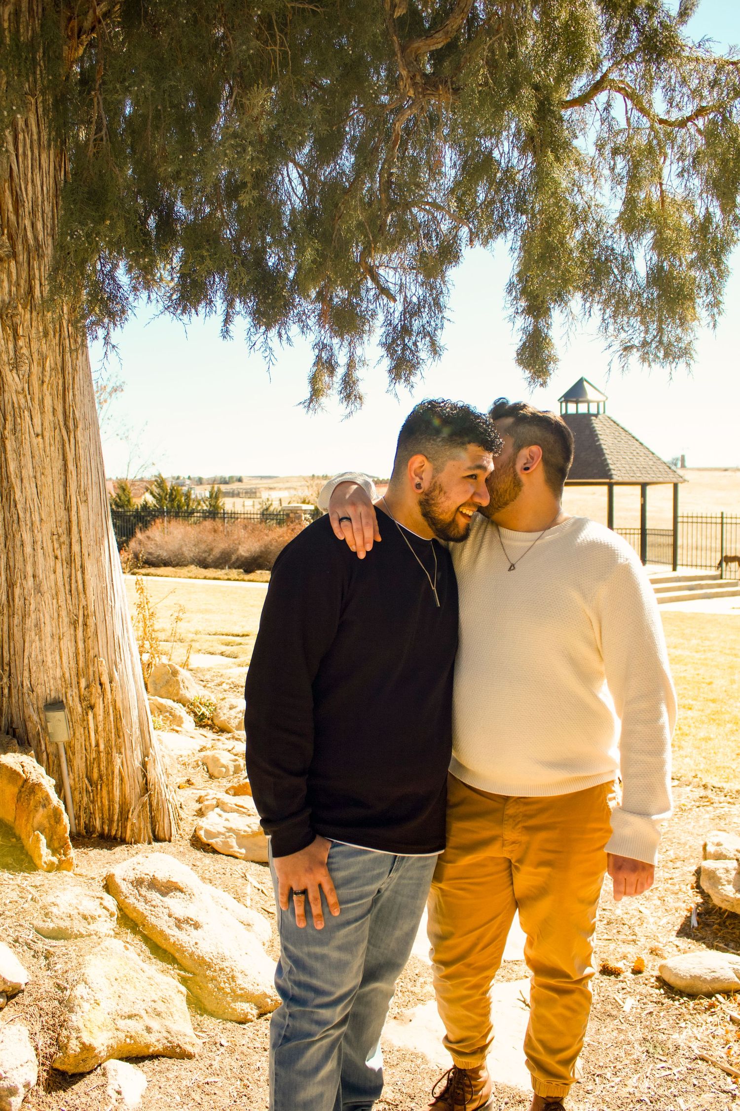 A couple shares an intimate moment under a large willow tree near a gazebo.