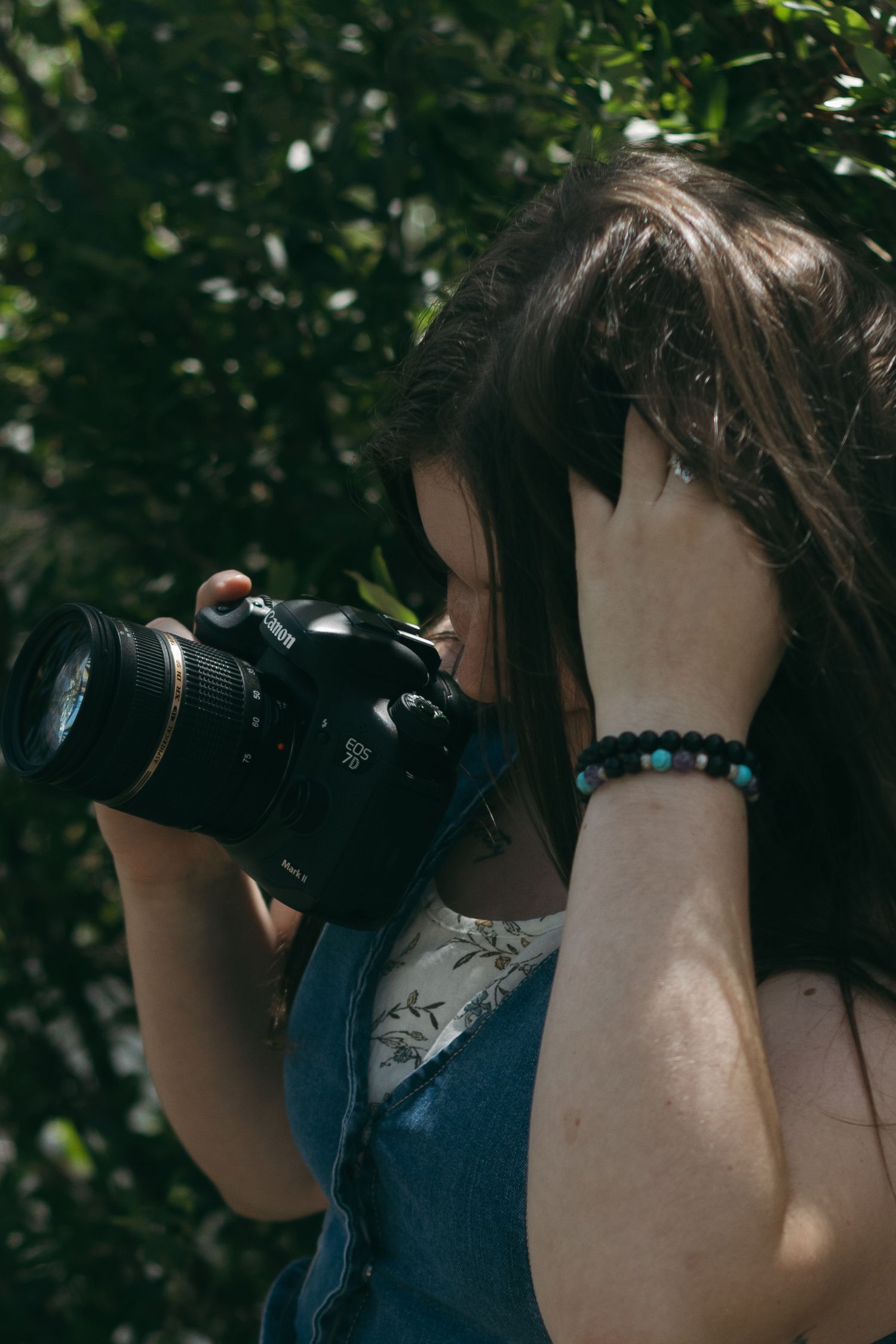 A person with dark hair uses a professional camera outdoors with green foliage in the background.